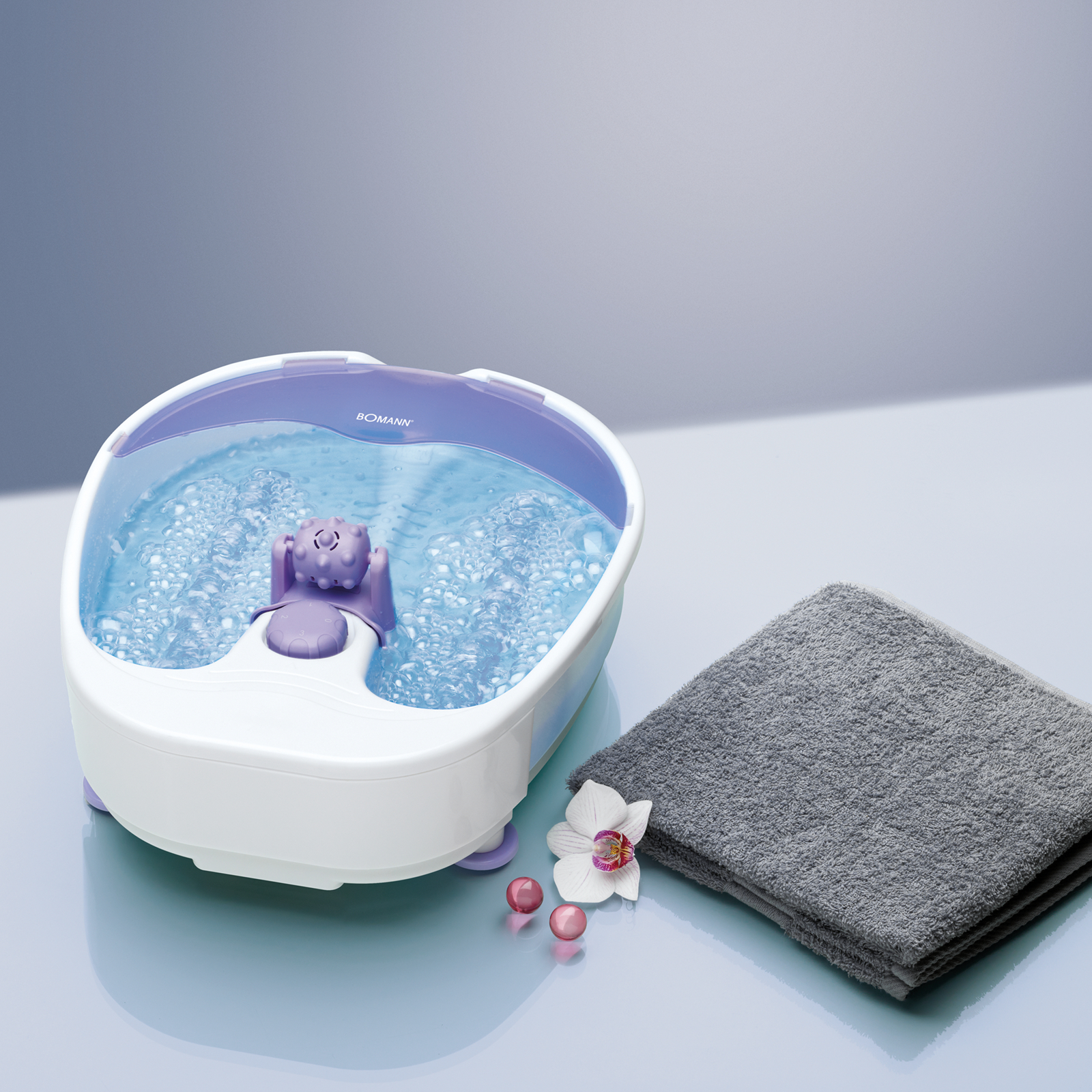 Ein Fußbad mit blauem Wasser und Blasen, eine lila Figur, ein graues Handtuch und eine Blume.