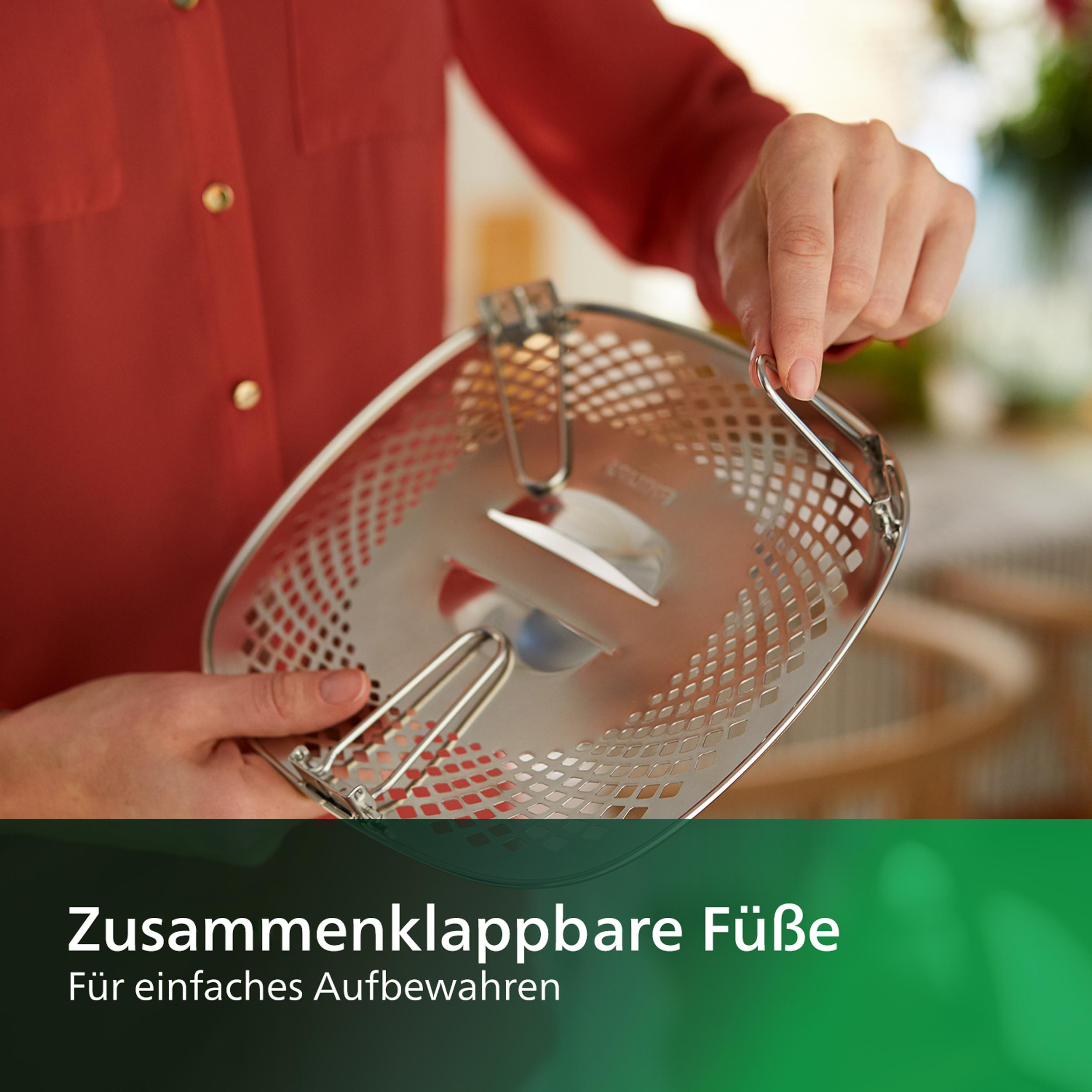 Eine Person hält einen silbernen Kochkorb mit klappbaren Griffen. Rotes Hemd im Hintergrund.