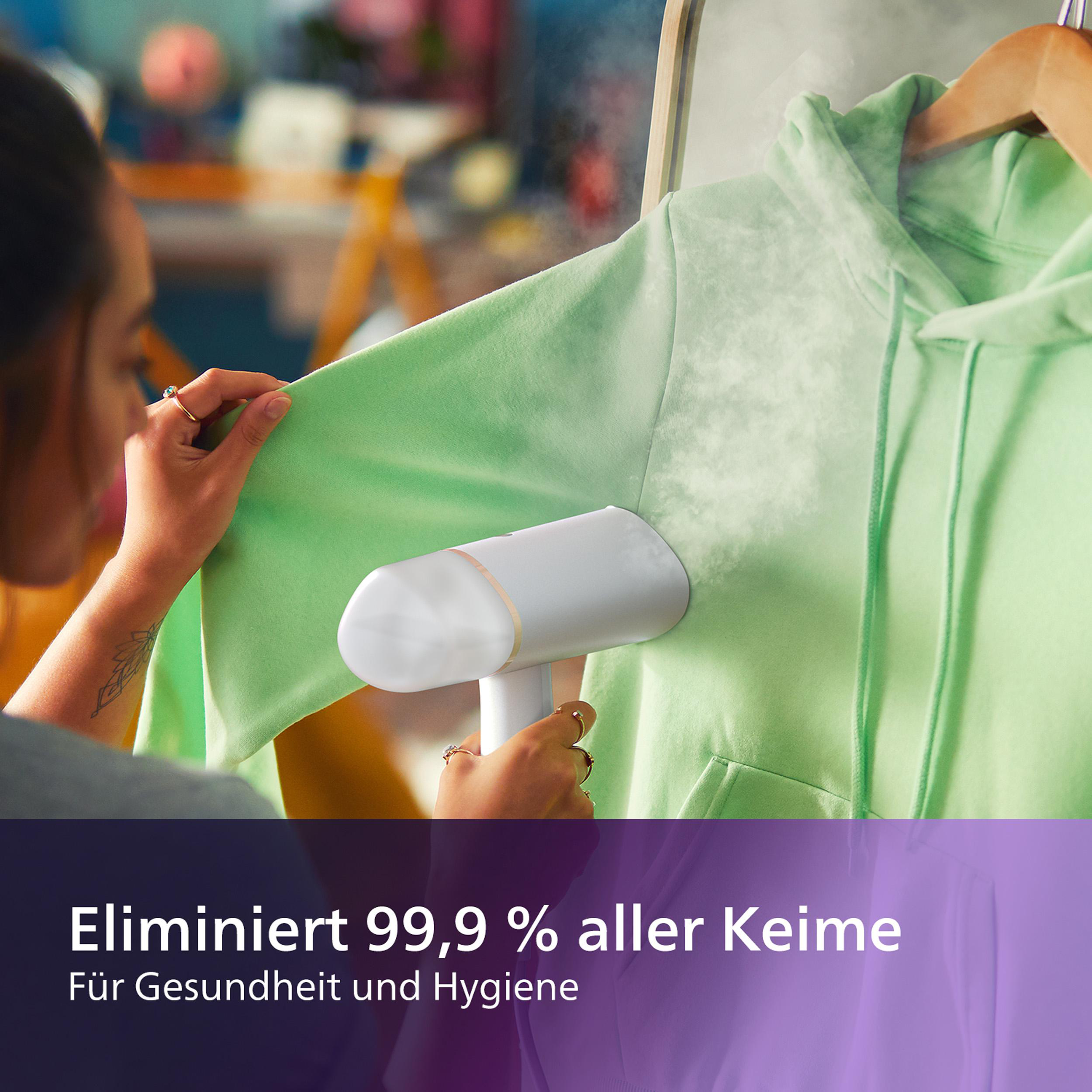 Eine Frau dämpft einen grünen Hoodie mit einem Handdampfer; Text: Eliminiert 99,9 % aller Keime.