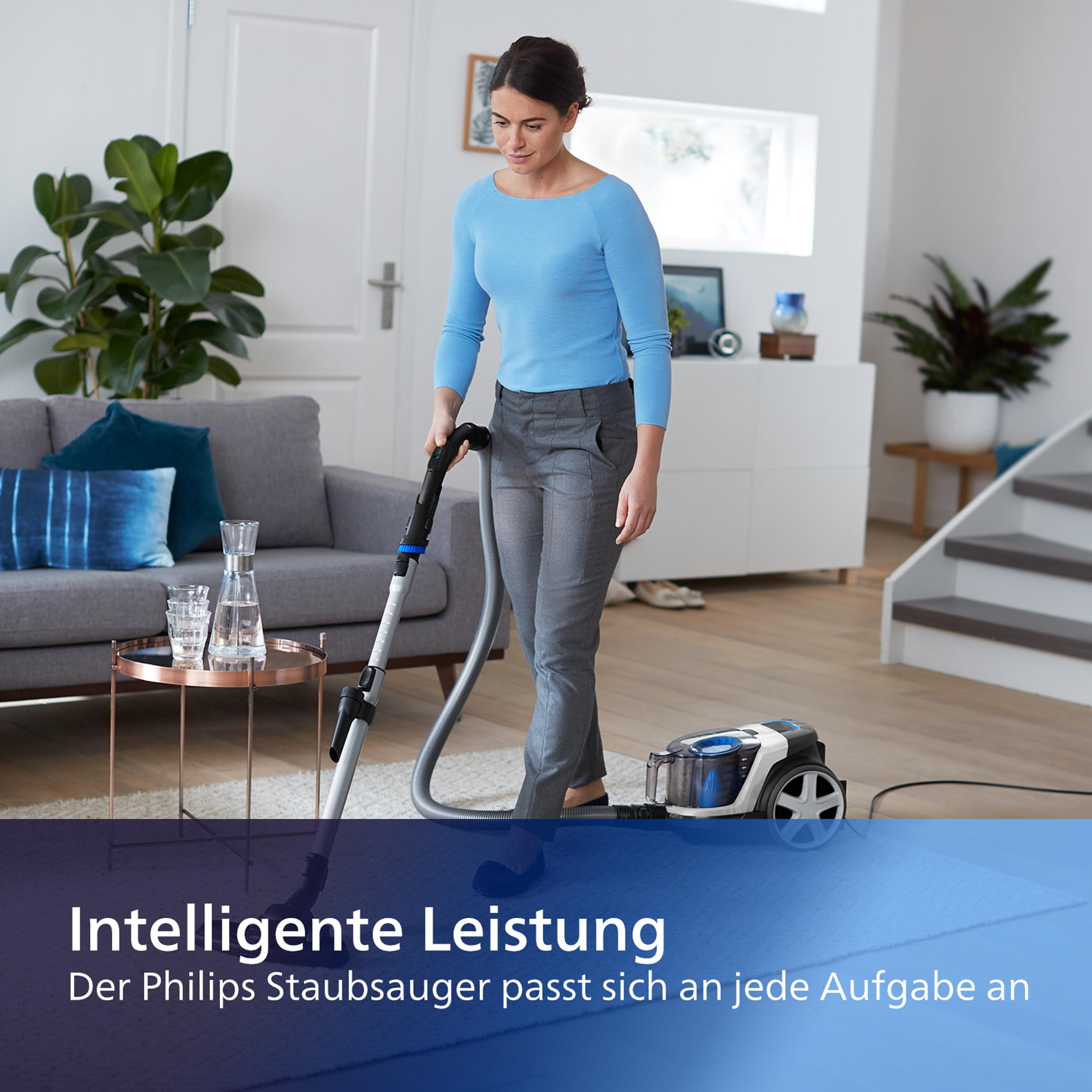 Eine Frau saugt das Wohnzimmer. Der Staubsauger ist schwarz und grau. Sofa, Teppich und Pflanzen sind im Hintergrund.