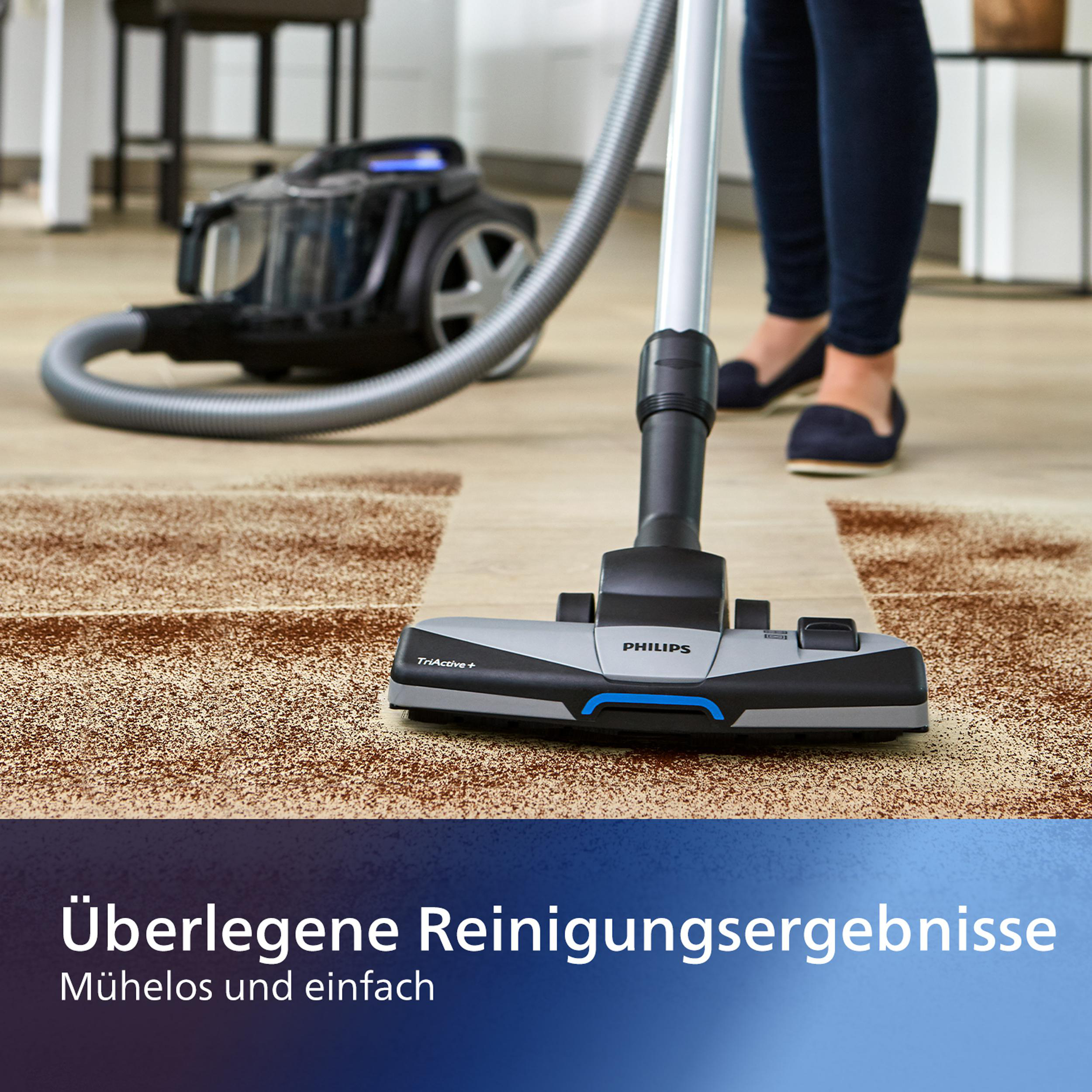 Ein Staubsaugerkopf auf einem braunen Teppich, mit einem Staubsauger im Hintergrund. Beine einer Frau sind sichtbar.