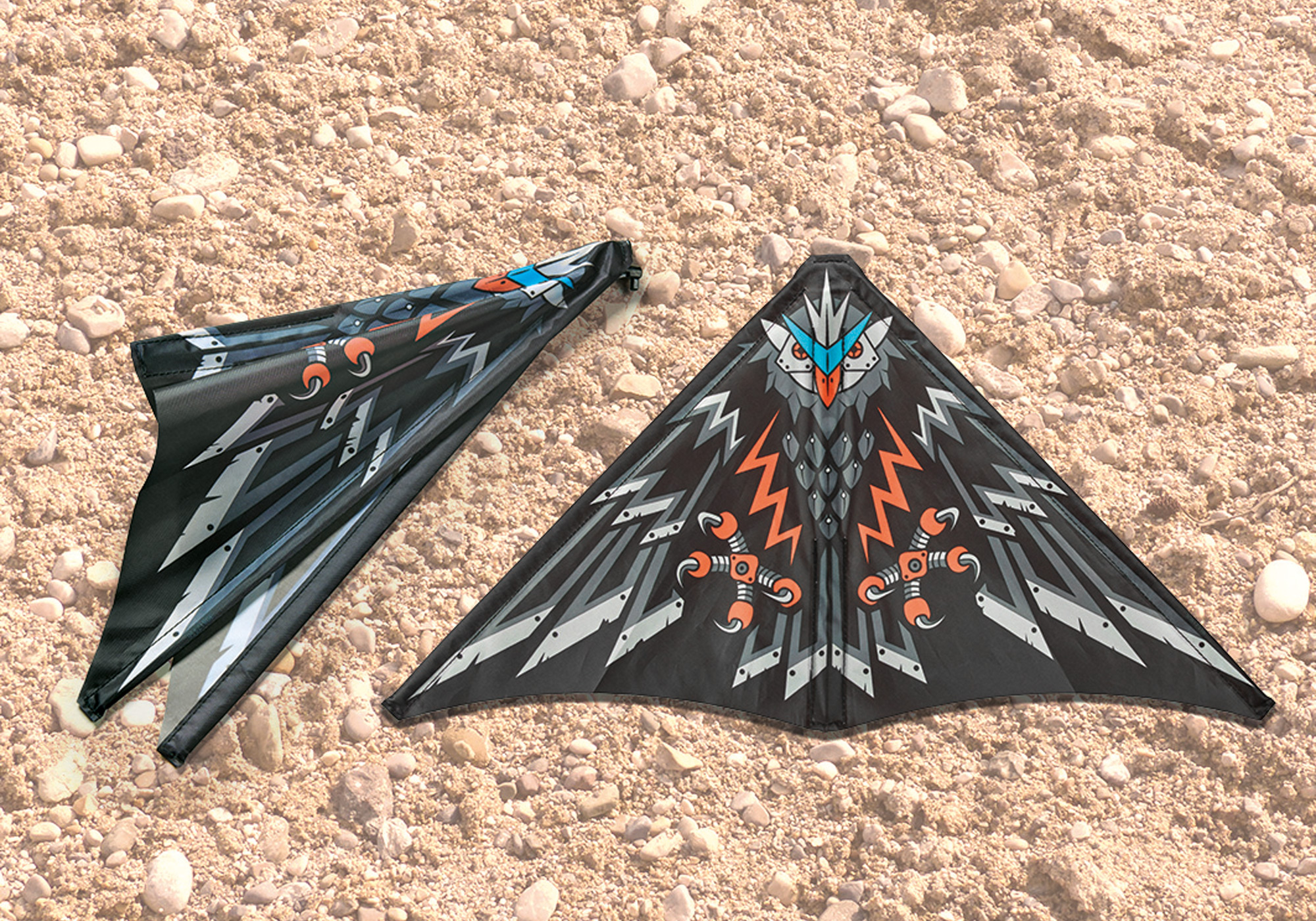 Zwei schwarze Drachen mit Vogelmotiven liegen auf einer strukturierten, sandigen Oberfläche.