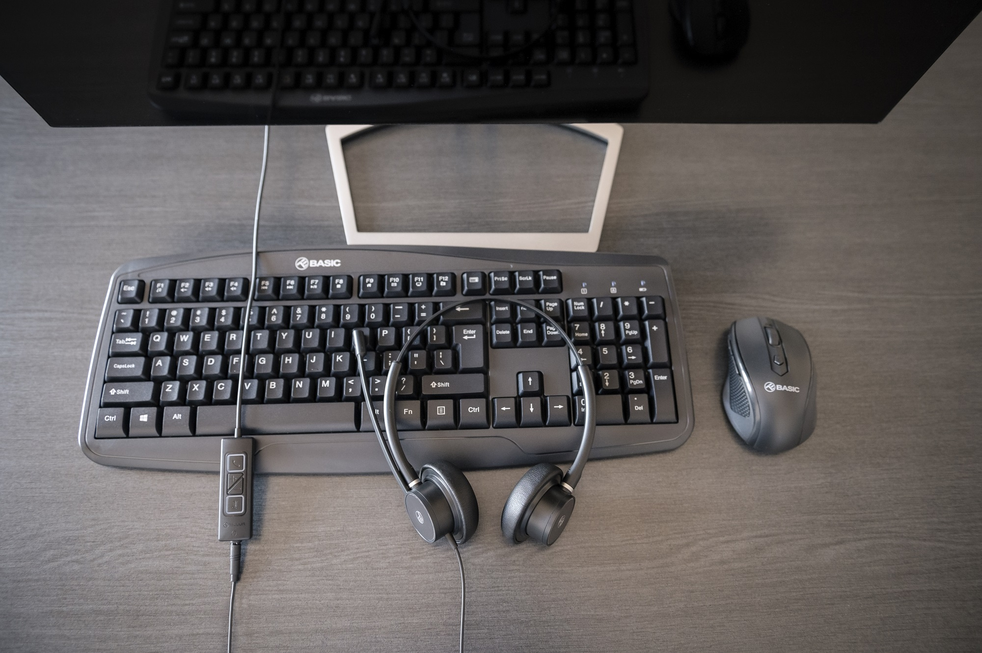 Konfiguracja biurka z klawiaturą, myszą, zestawem słuchawkowym i monitorem. Szara powierzchnia imitująca drewno.