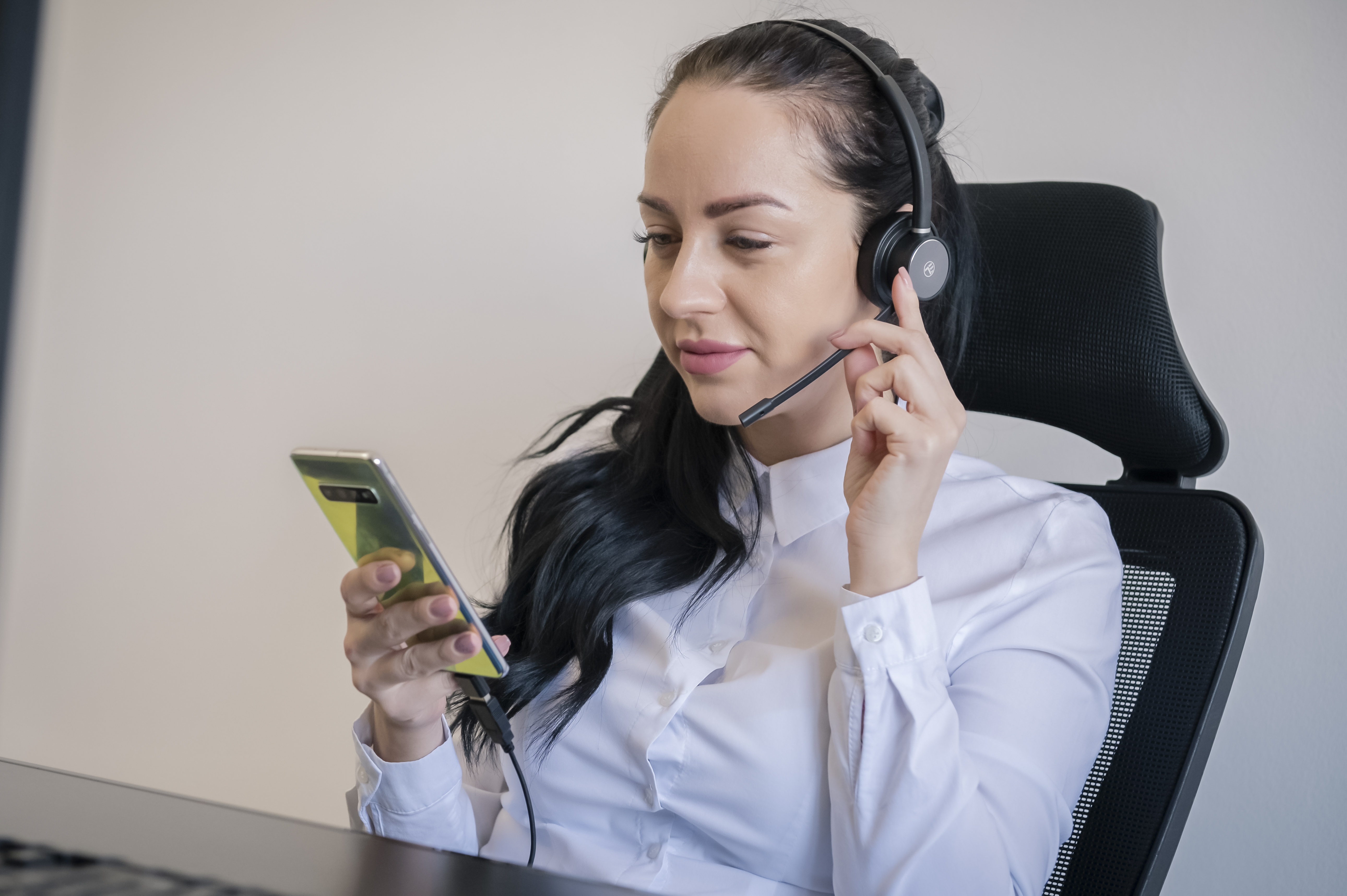 Kobieta z zestawem słuchawkowym i telefonem, siedząca przy biurku. Trzyma telefon i rozmawia.
