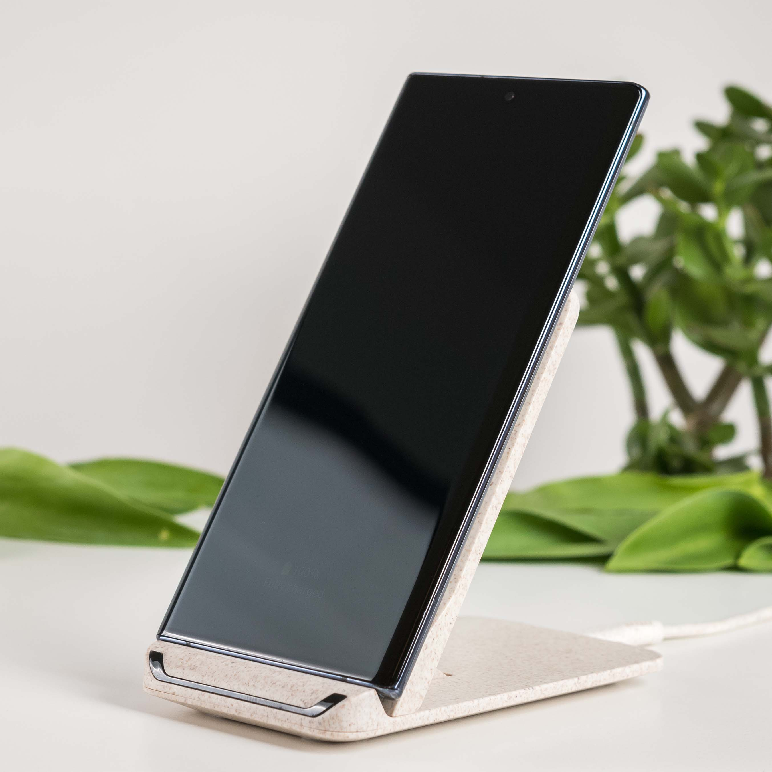 Czarny telefon na beżowej bezprzewodowej podstawce ładującej, z zieloną rośliną w tle.