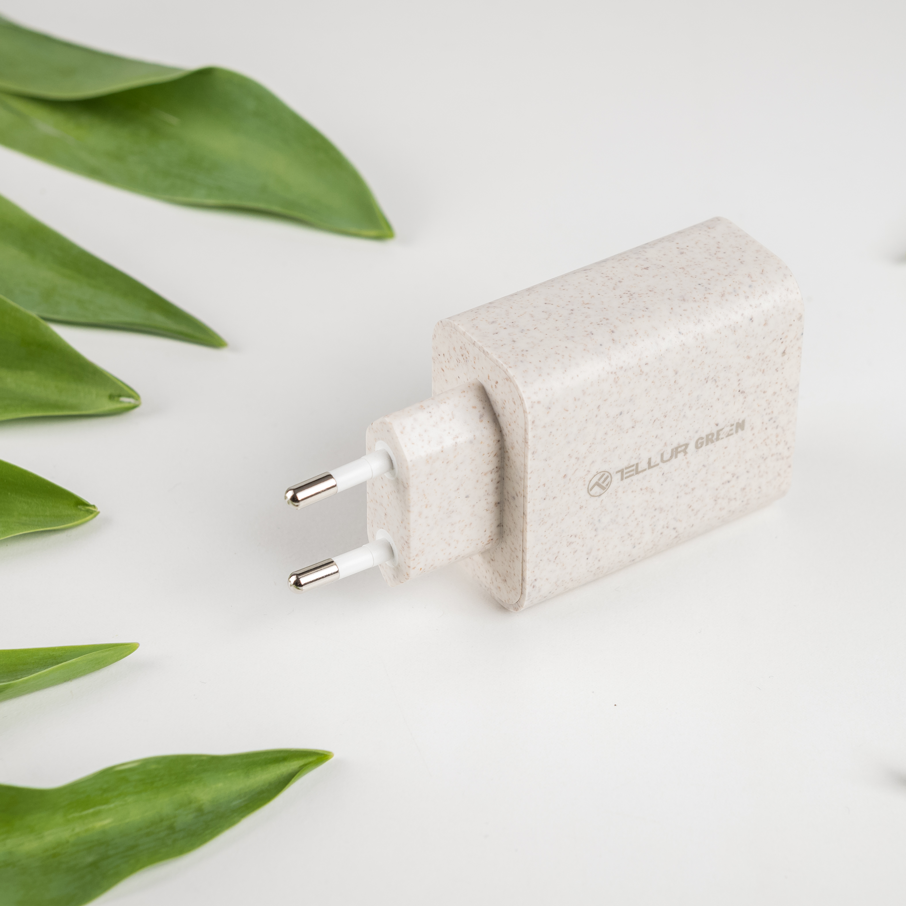 Beżowa ładowarka z dwoma bolcami i logo, otoczona zielonymi liśćmi na białym tle.
