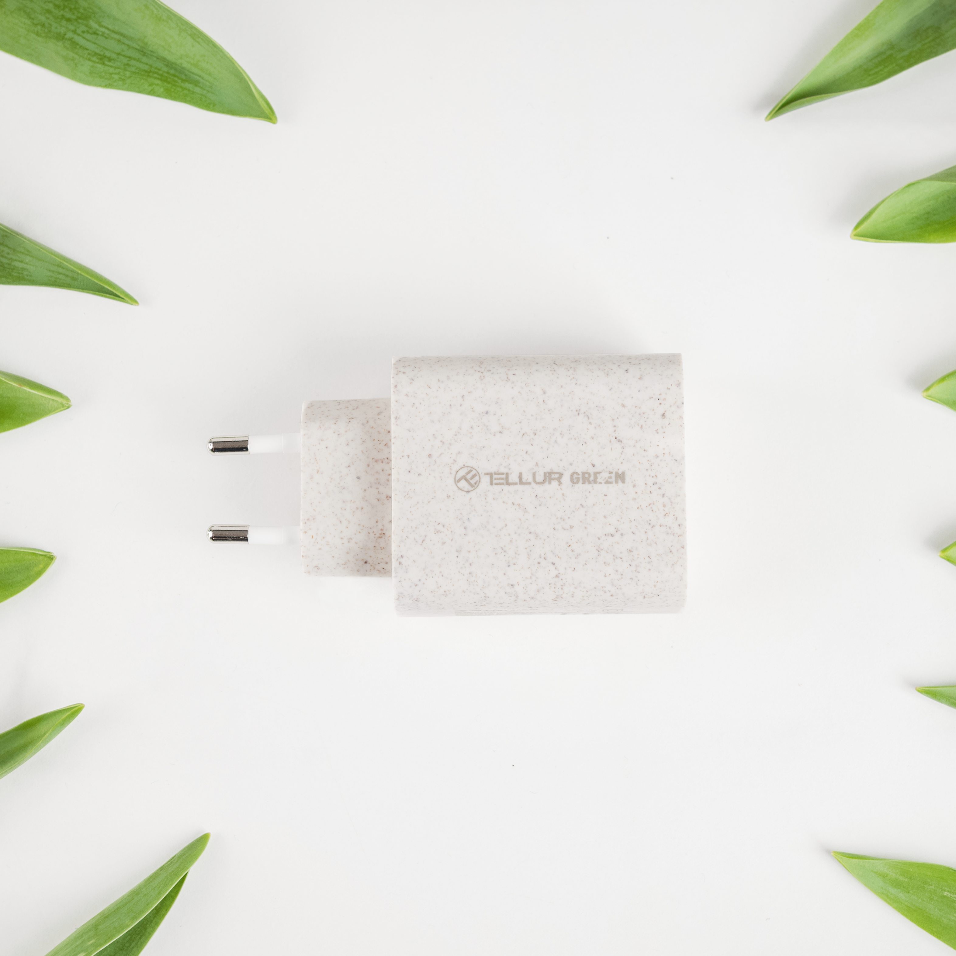 Biała ładowarka z logo 'Tellur Green', otoczona zielonymi liśćmi na białej powierzchni.