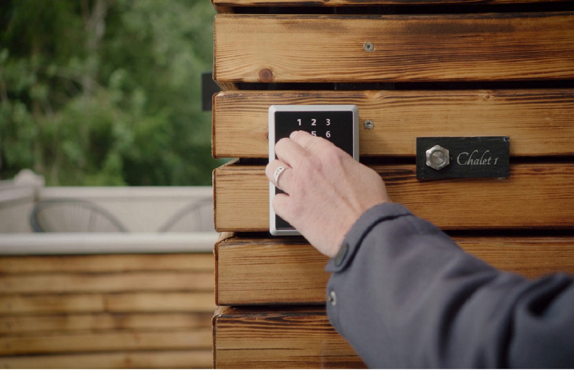 Die Hand einer Person gibt einen Code auf einer Tastatur ein, um ein Holztor mit einem Chalet-Schild zu öffnen.