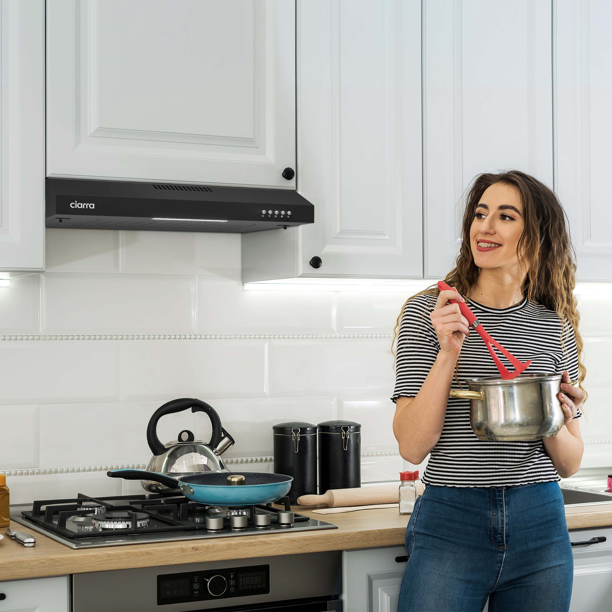Femme dans la cuisine tenant une casserole et un fouet rouge, regardant à droite.