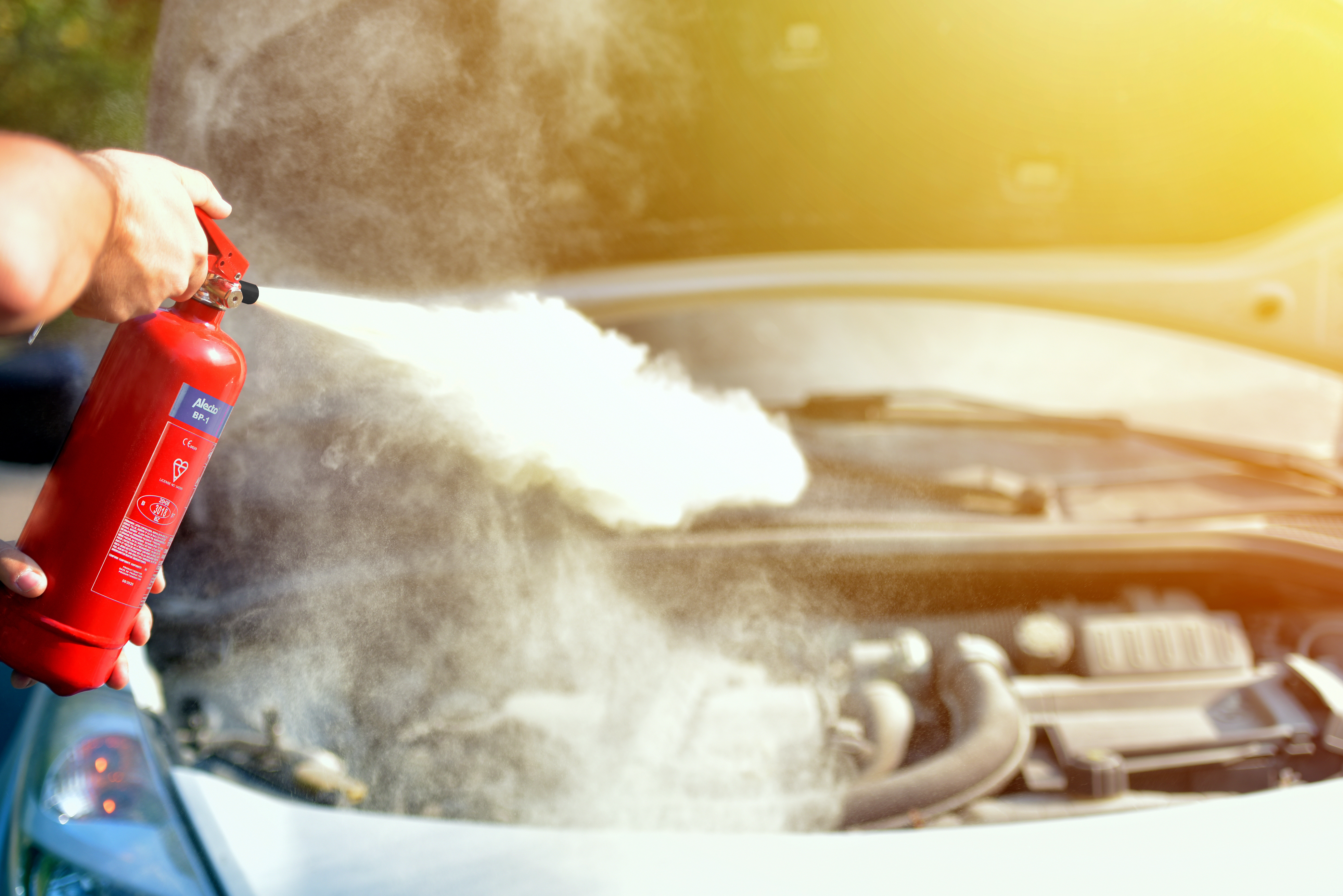 A person is spraying a red fire extinguisher on a car engine, smoke is visible.