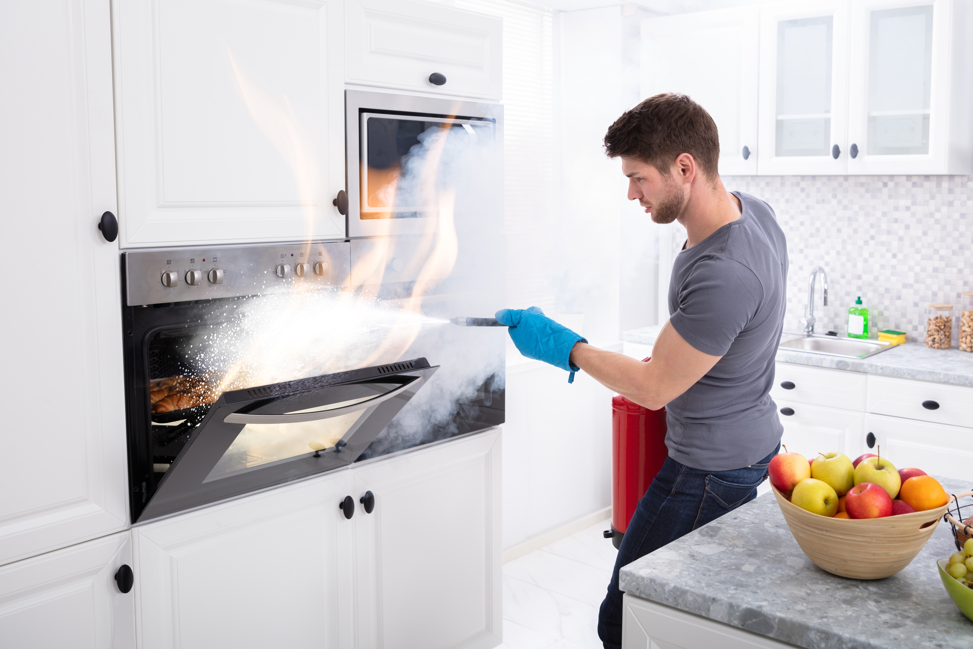 Man in kitchen extinguishing oven fire with fire extinguisher.