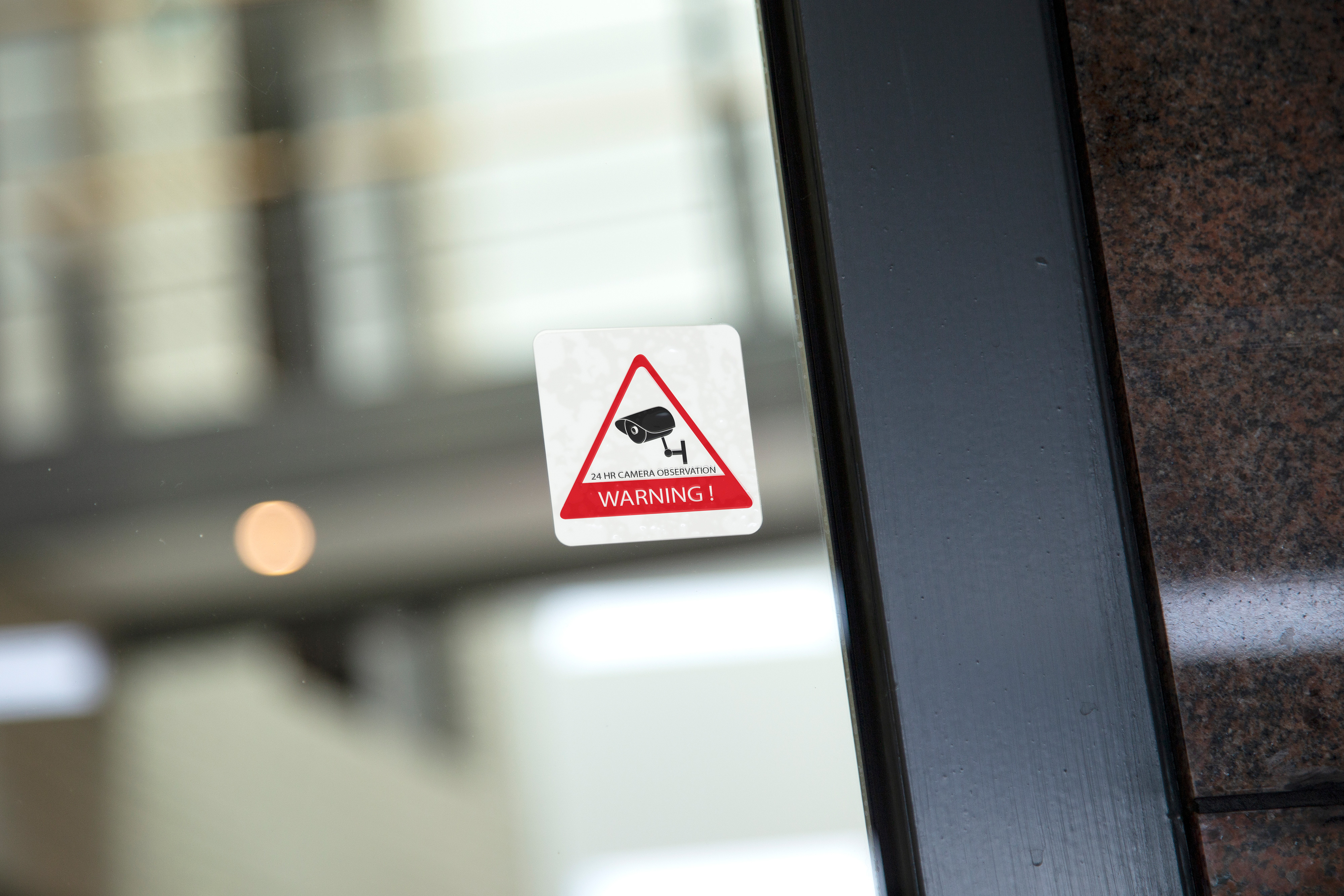 Warning sticker on a glass door. Red triangle with camera icon and text. Blurred background.