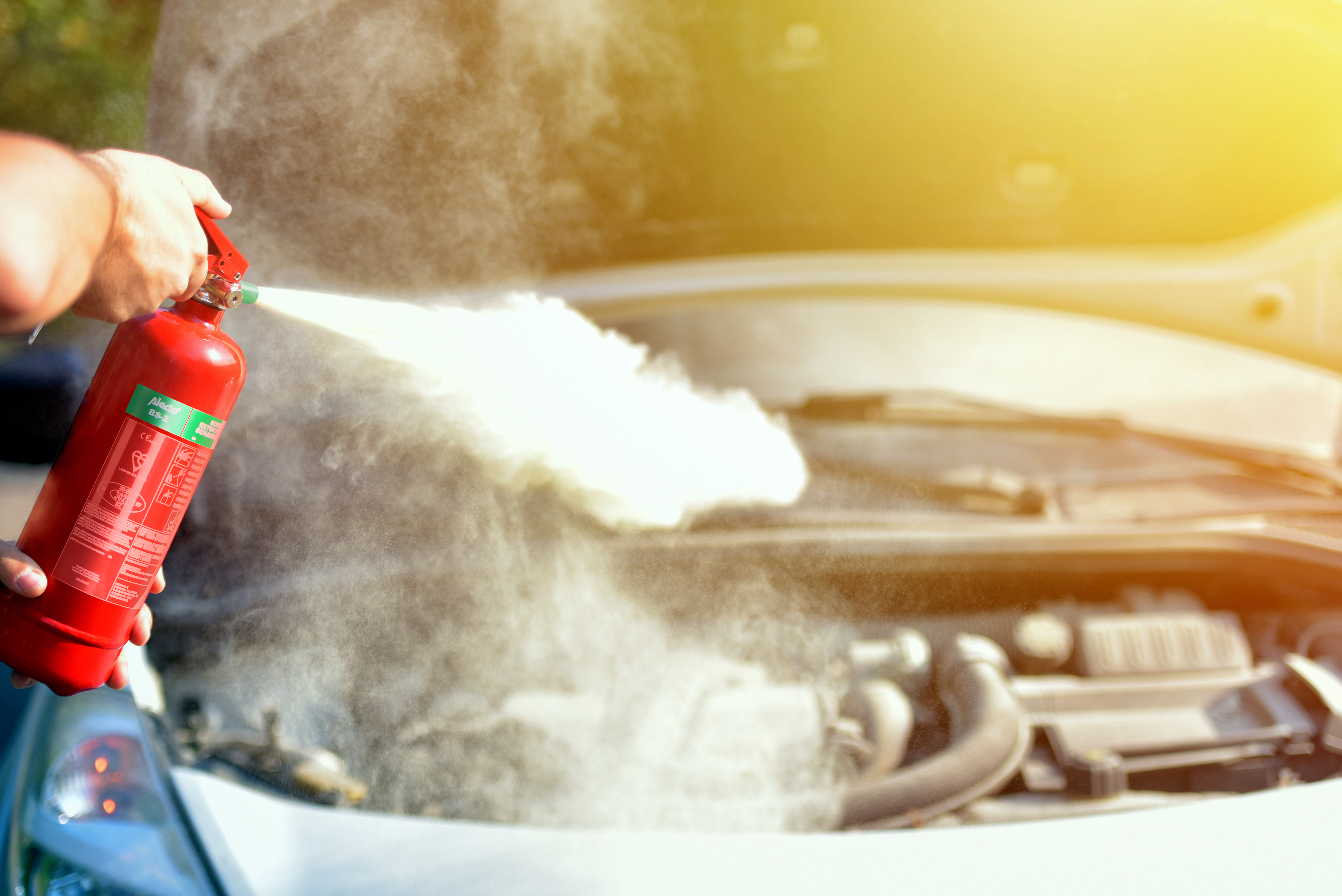Hand spraying a red fire extinguisher on a car engine, with smoke and sunlight.
