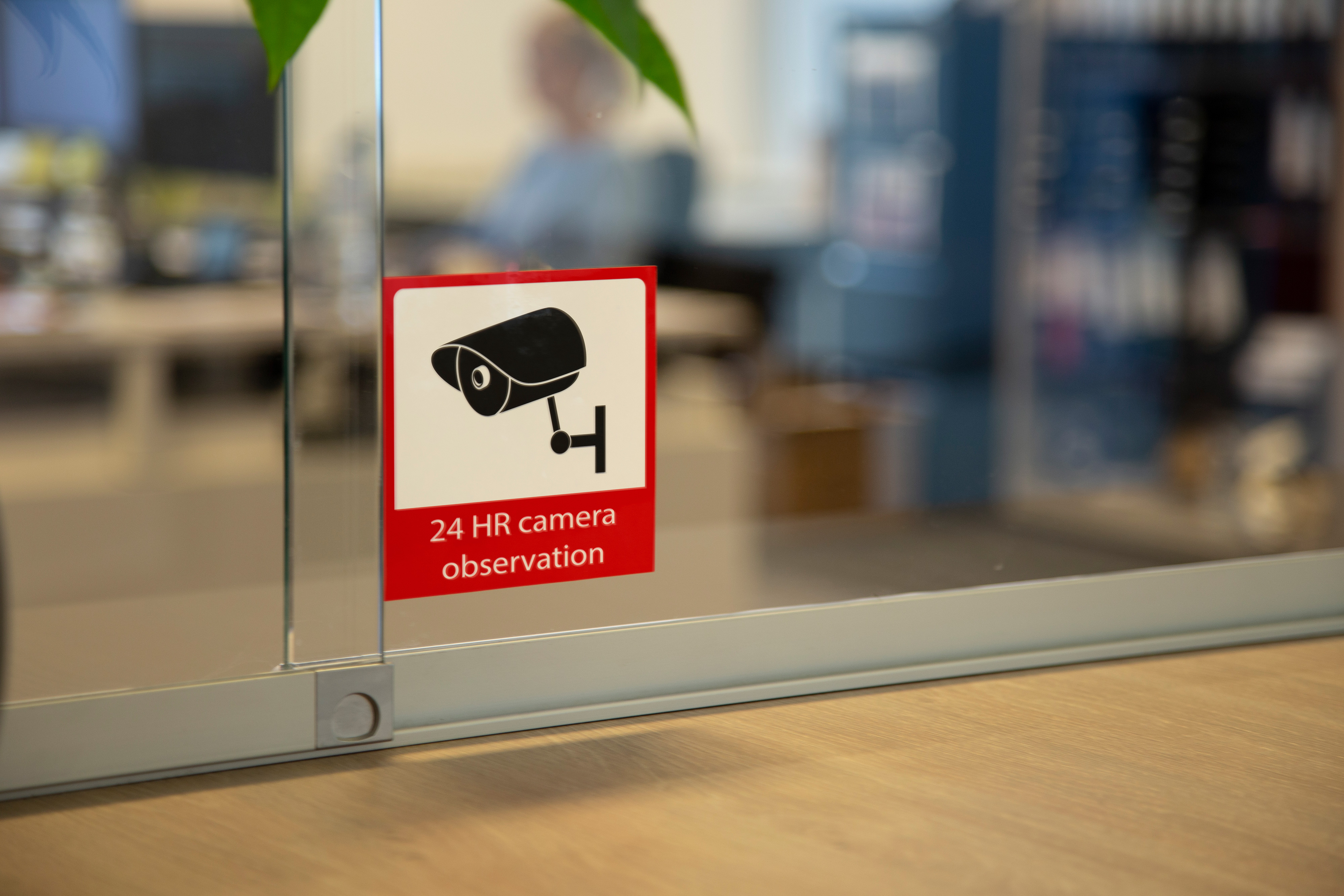 A sign on glass reads '24 HR camera observation' with a camera icon. A person is blurred in the background.