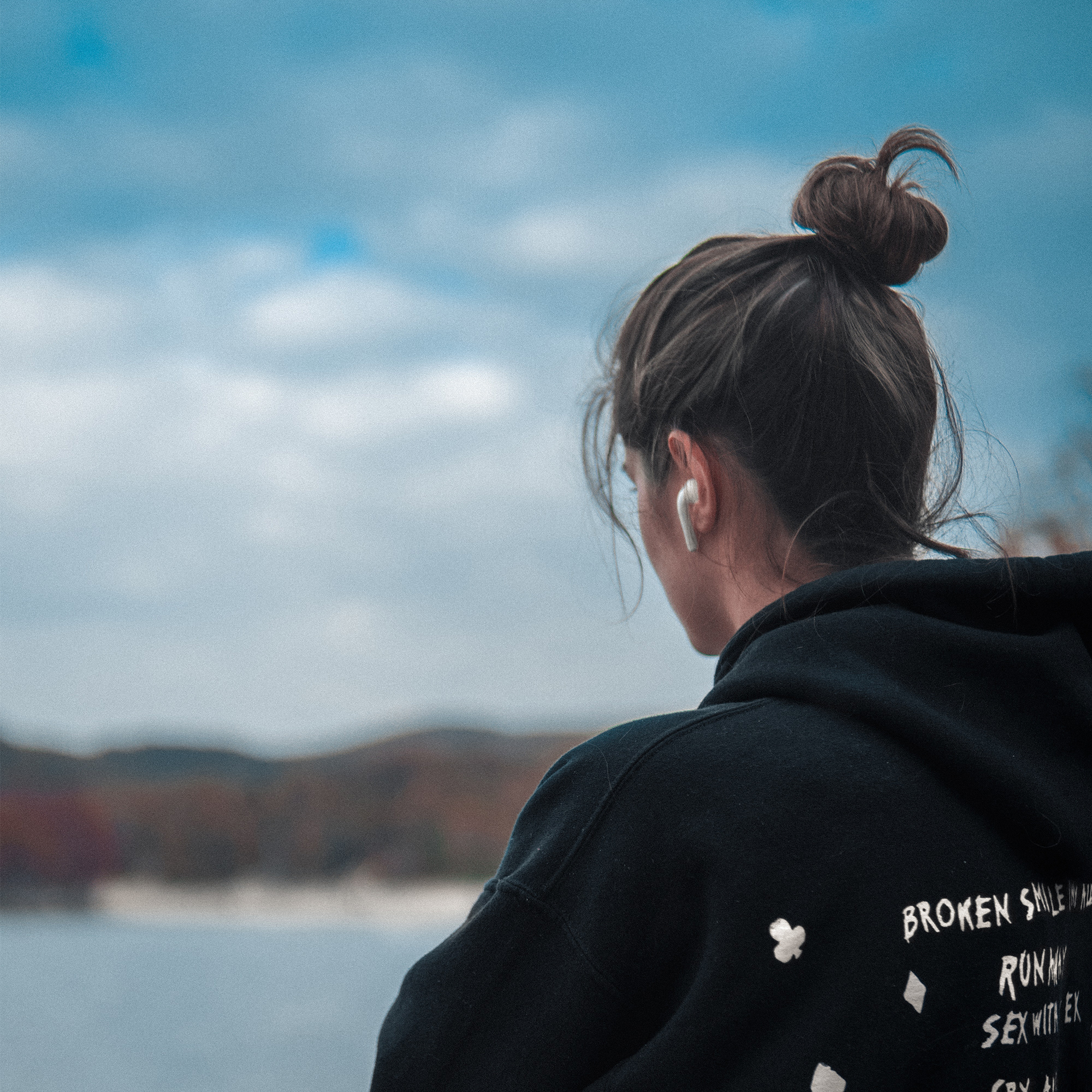 Eine Frau mit Dutt und Ohrhörern blickt auf einen See mit bewölktem Himmel.