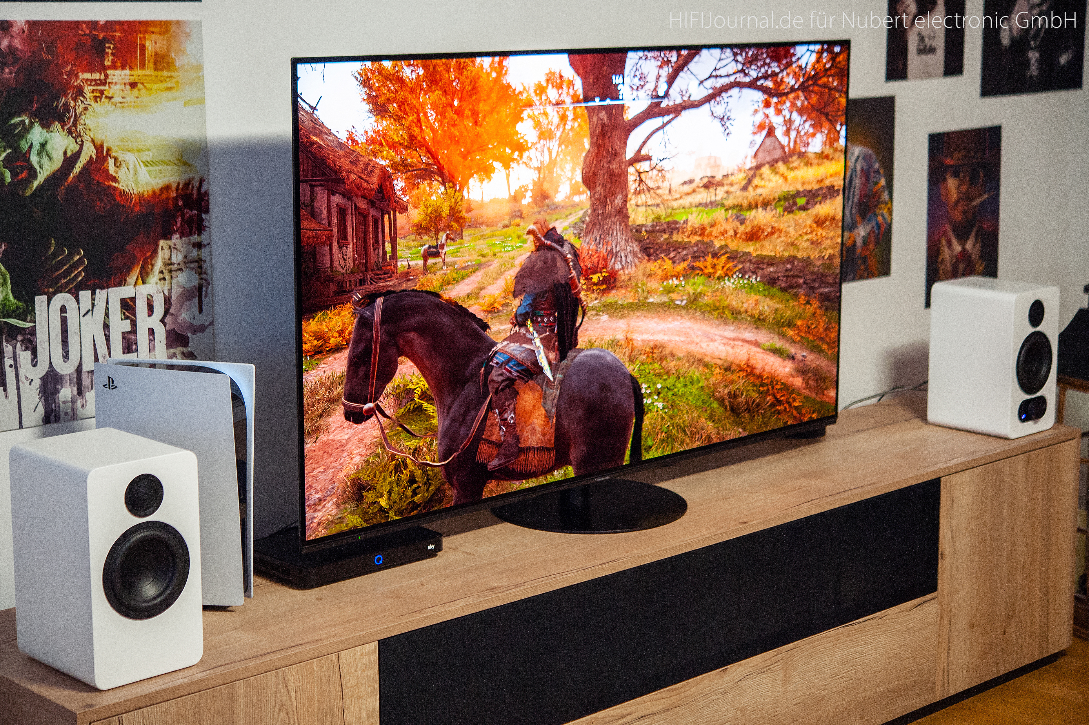 Ein Fernseher, der eine Spielszene anzeigt, flankiert von weißen Lautsprechern und einer PlayStation. Holztisch.