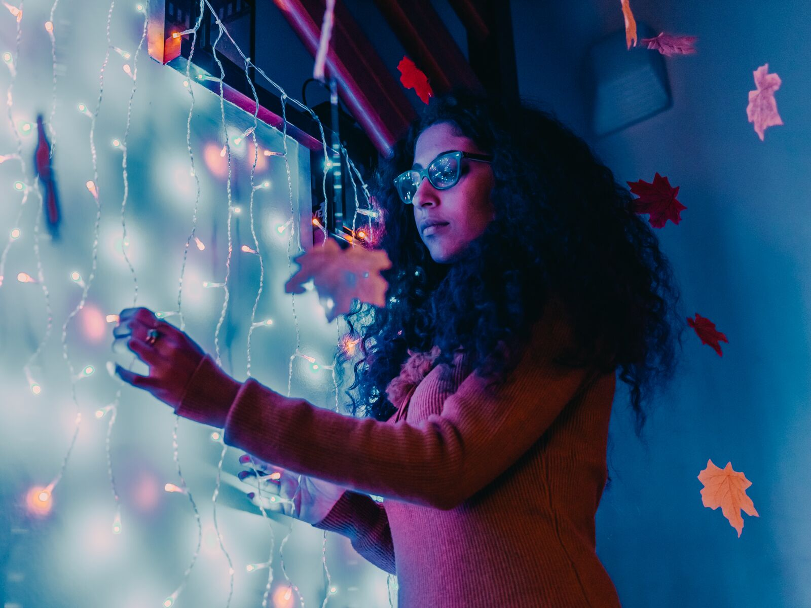 Kobieta w okularach sięgająca po wiszące liście z światłami. Miękkie oświetlenie, niebieskie i czerwone odcienie.