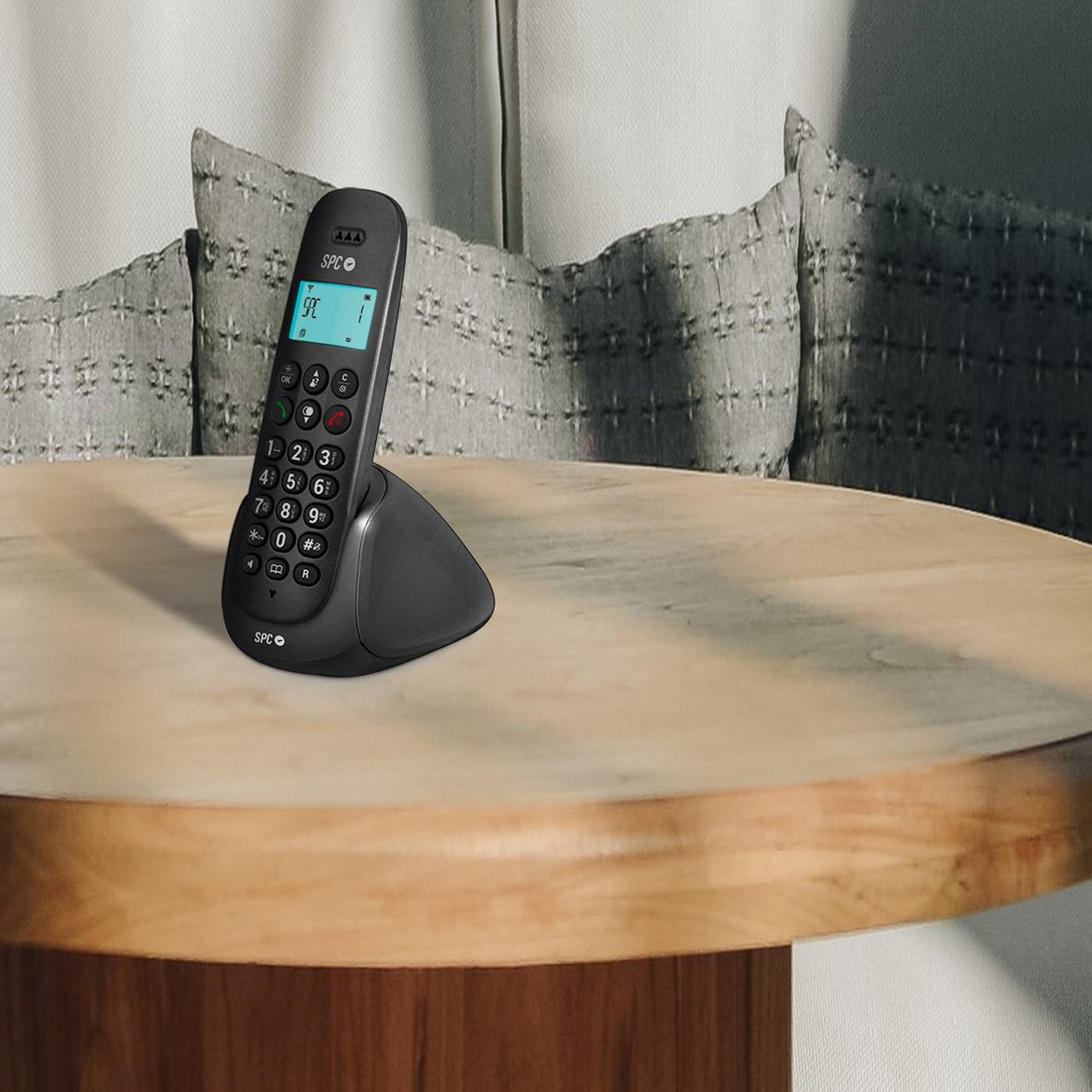Un teléfono fijo negro sobre una mesa de madera. Hay un fondo con almohadas y una pared.