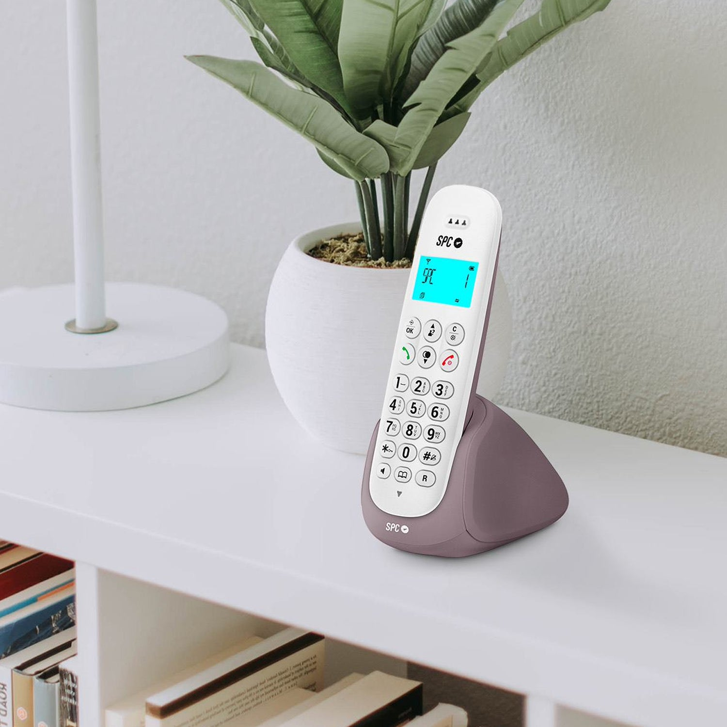 Teléfono inalámbrico en una estantería. El teléfono y la base son blancos y morados. Planta en el fondo.