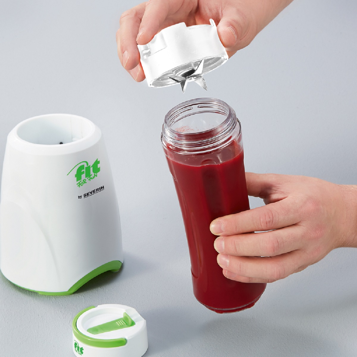 Handen die een blender monteren met rode vloeistof en een witte blenderbasis met groene accenten.
