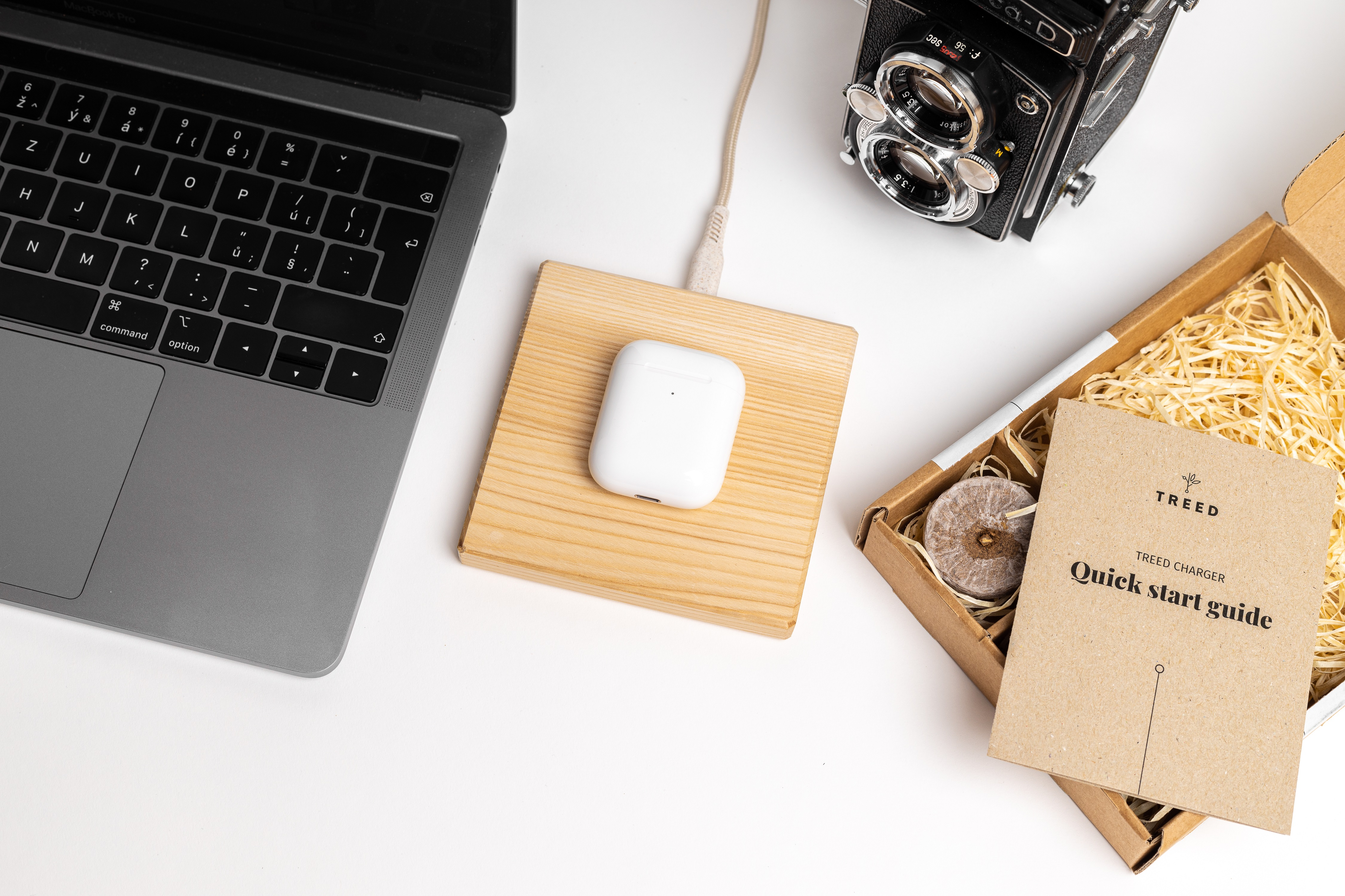 Laptop, Airpods, Kamera und eine Schnellstartanleitung auf einem hölzernen Ladepad, weiße Oberfläche.