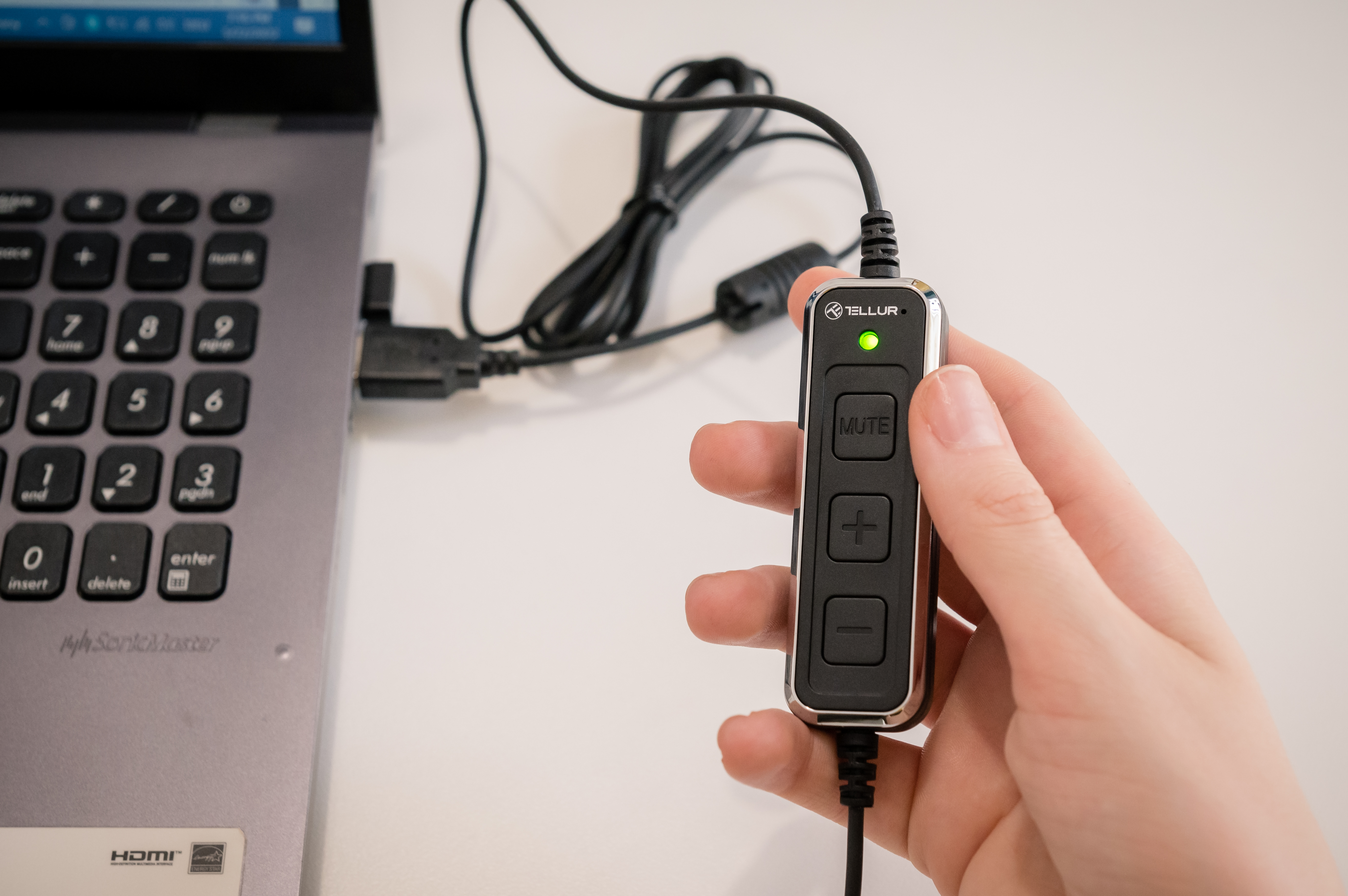 Dłoń trzymająca czarny dongle do sterowania audio z przyciskiem wyciszania, podłączony do laptopa.