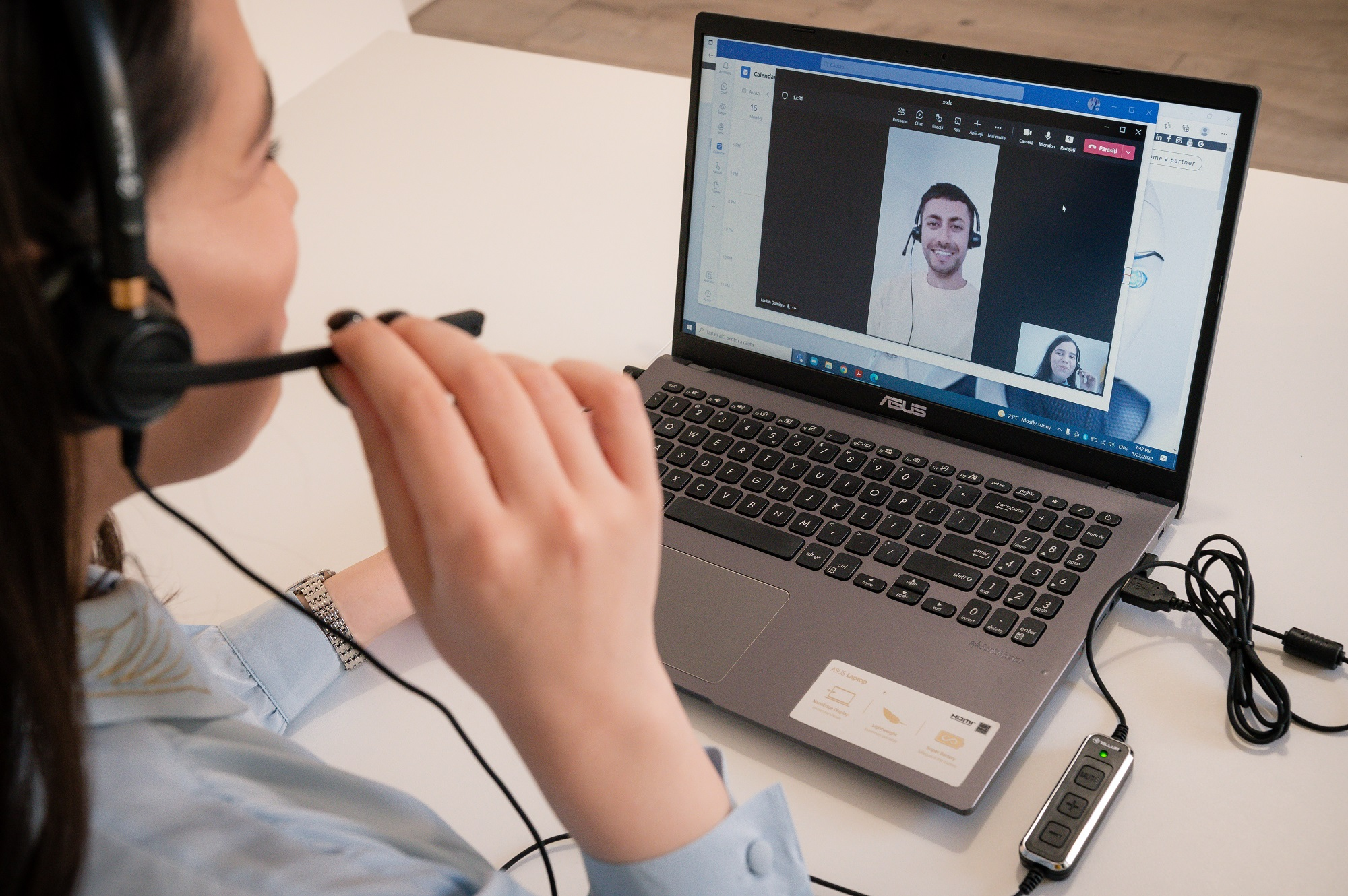 Kobieta w zestawie słuchawkowym prowadzi rozmowę wideo z dwiema osobami na ekranie laptopa.