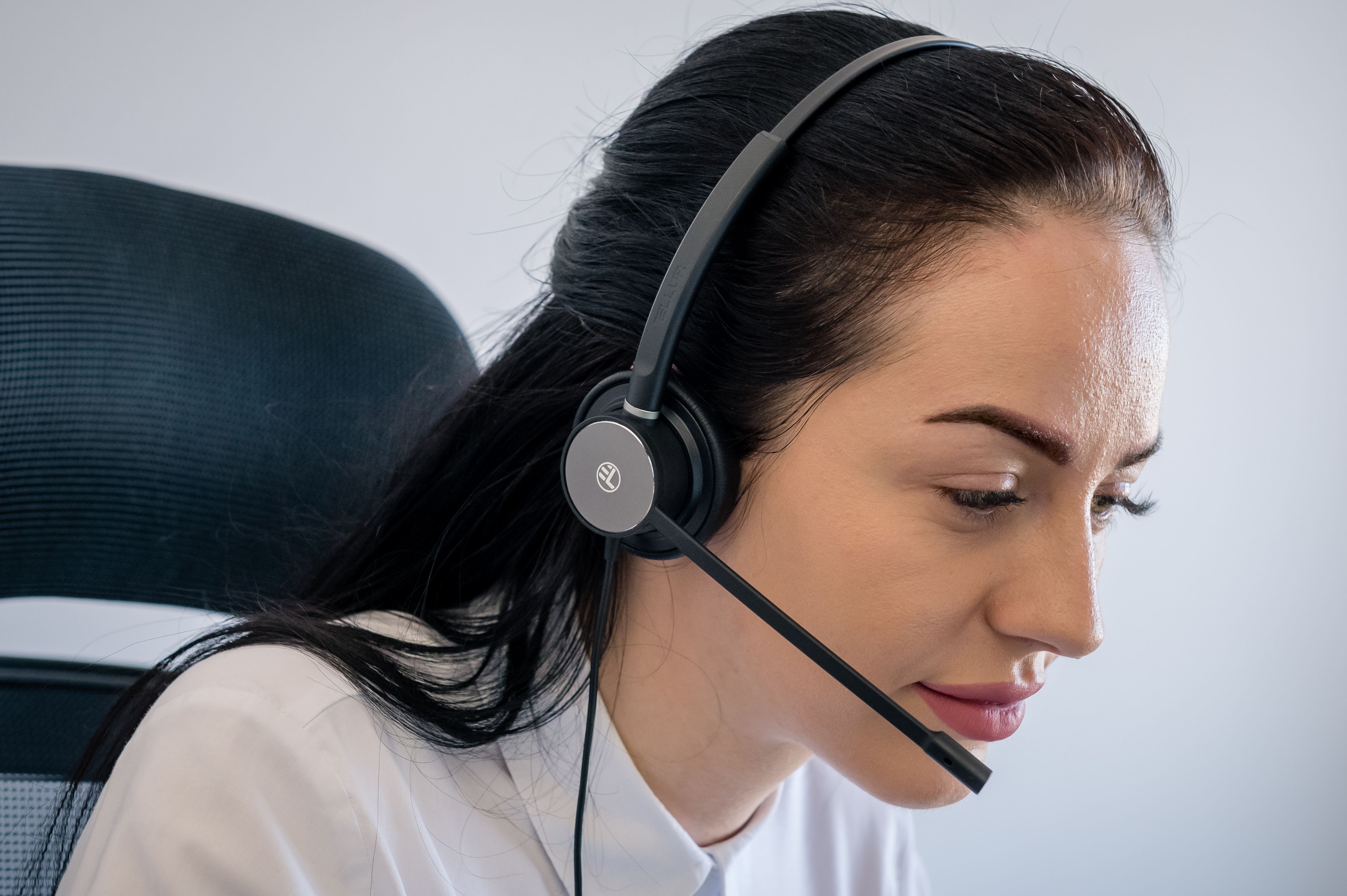 Kobieta w zestawie słuchawkowym. Patrzy w dół i przebywa w biurze. Biała koszula.