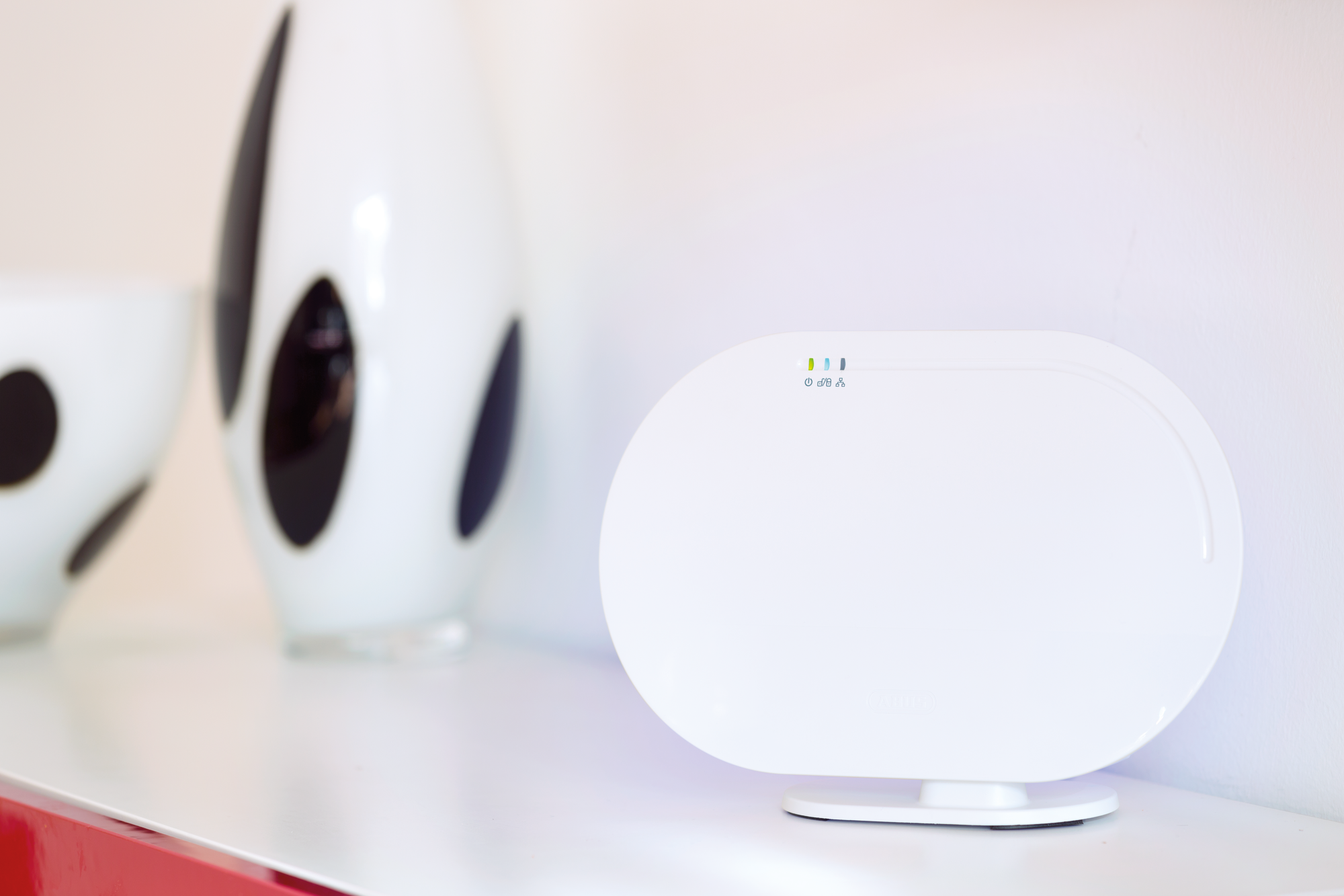 Weißes Smart-Home-Gerät auf einem weißen Regal, mit zwei dekorativen Vasen dahinter.