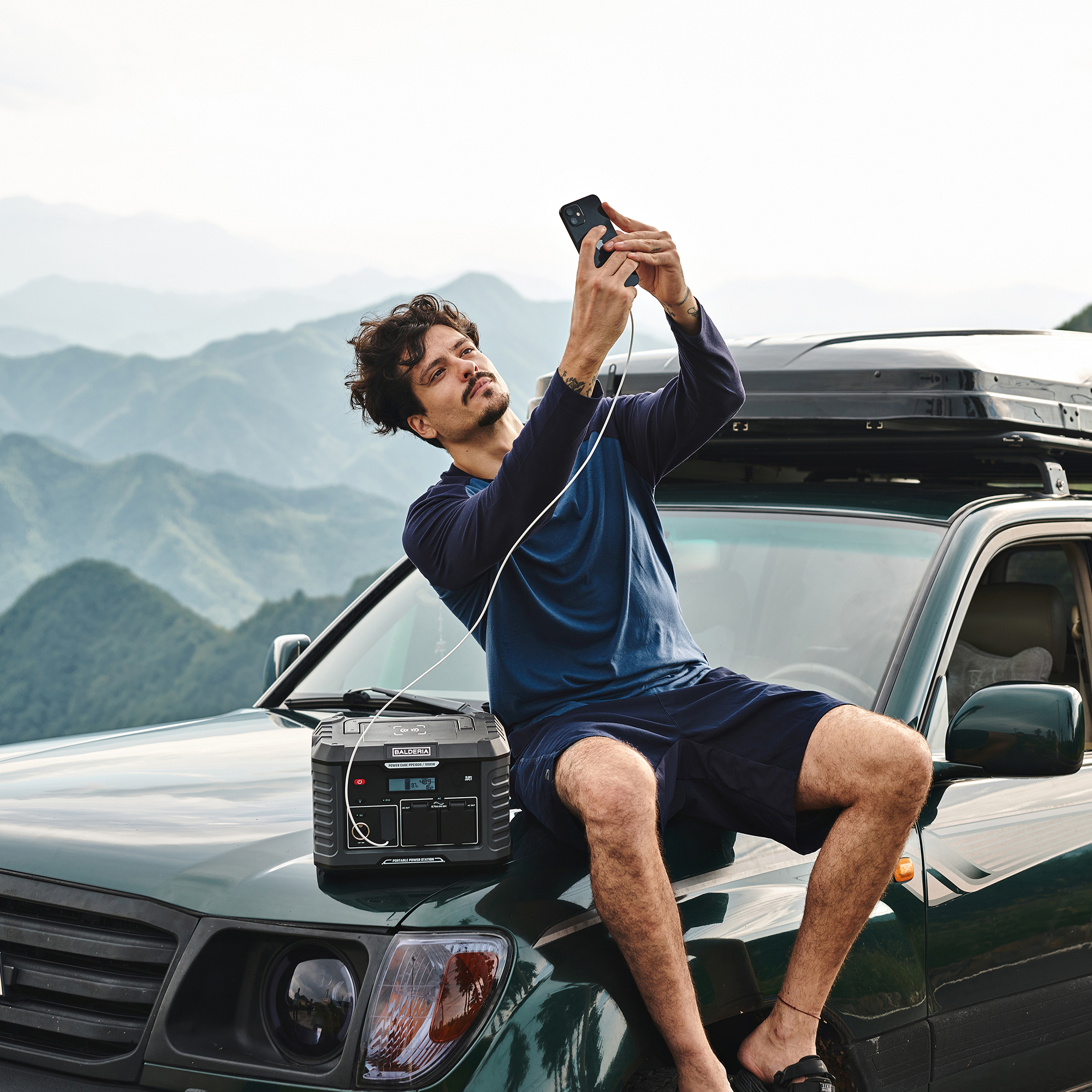 Mann macht Selfie auf Motorhaube, mit schwarzem Kraftwerk und Bergen im Hintergrund.