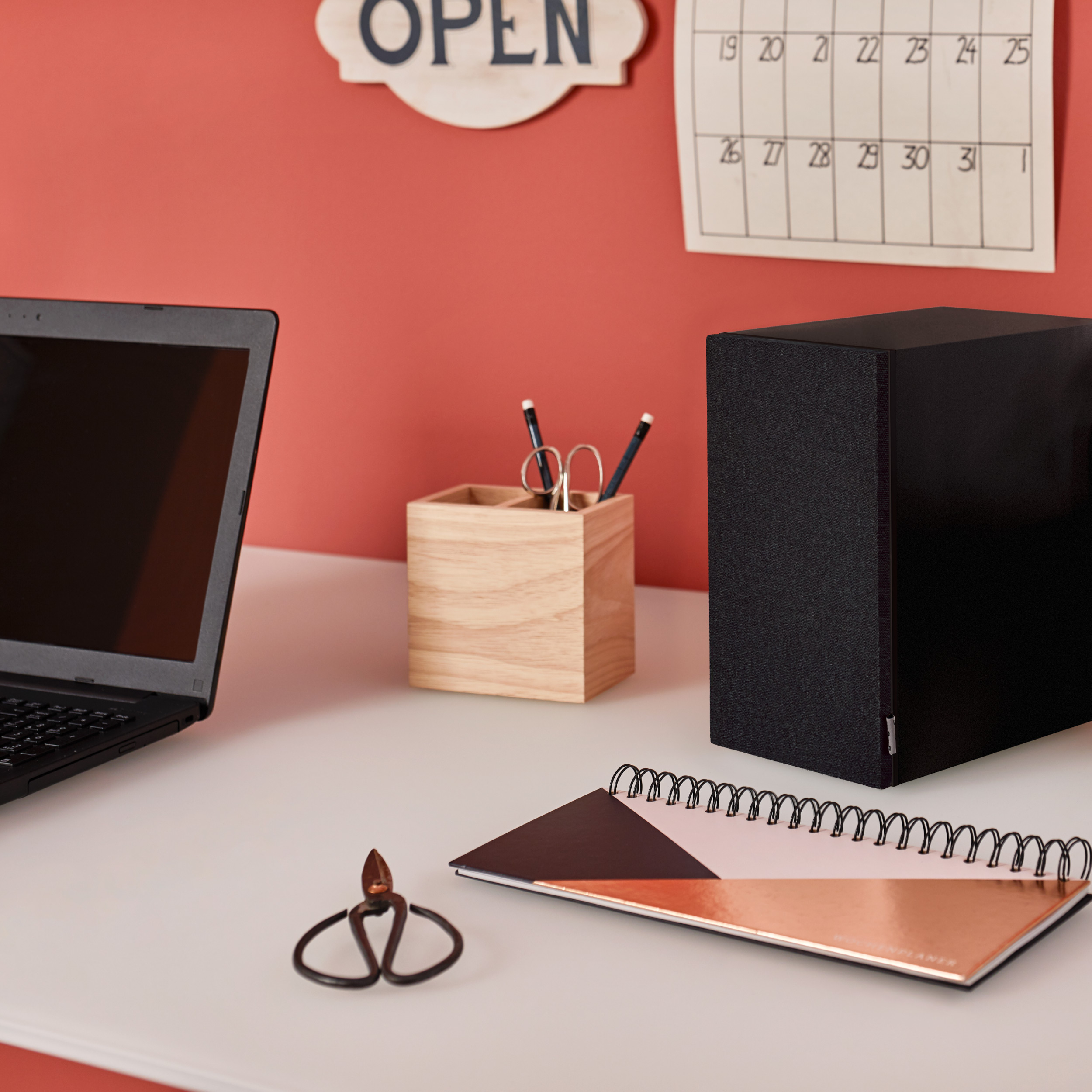 Büroszene mit Laptop, Lautsprecher, Notizbuch, Bleistiften und offenem Schild an roter Wand.