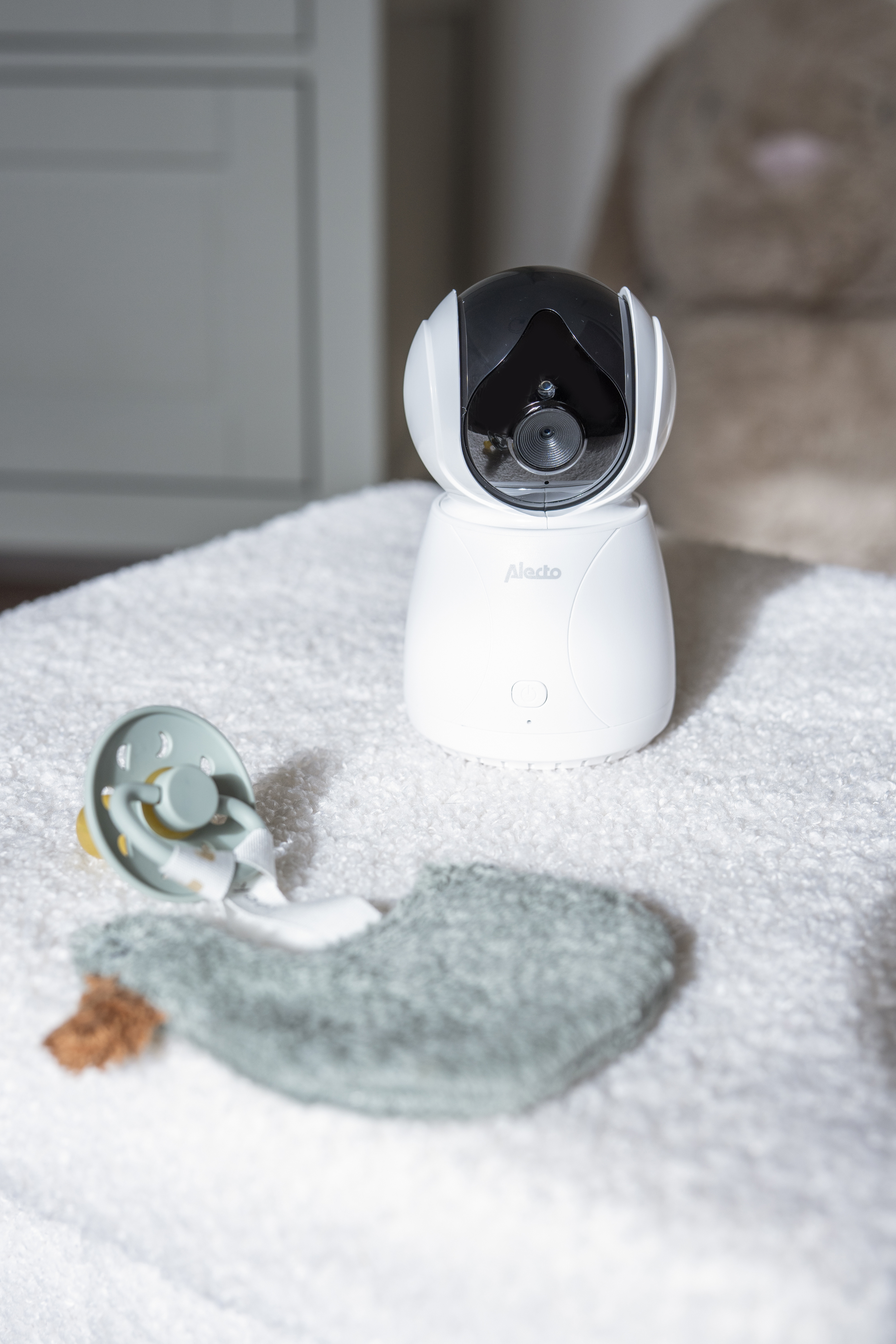 Weißer Babyphone mit schwarzem Oberteil, Schnuller und einem grünen Spucktuch auf weißem Handtuch, unscharfer Hintergrund.