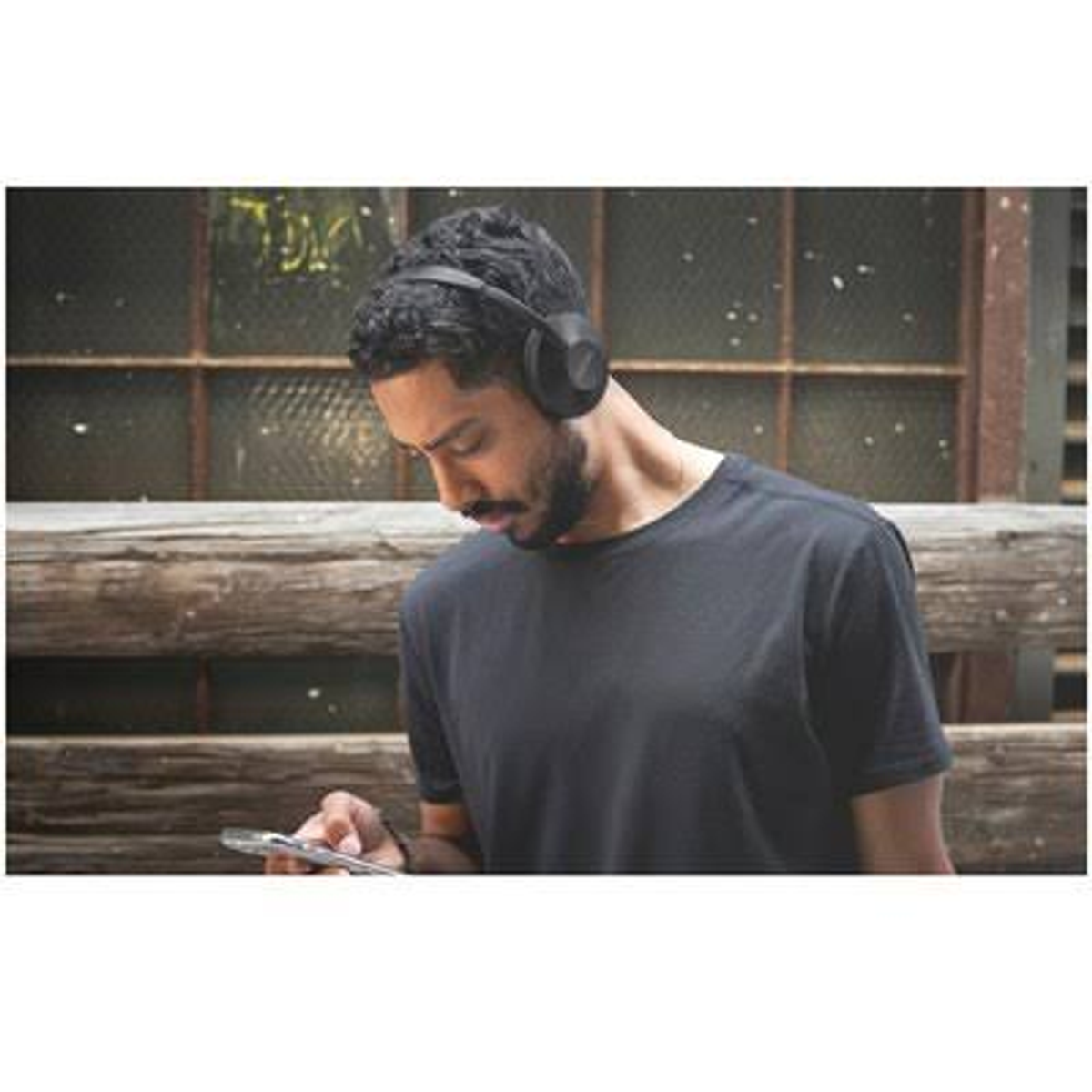 Un uomo con la barba con cuffie nere, tiene un telefono vicino a una finestra.