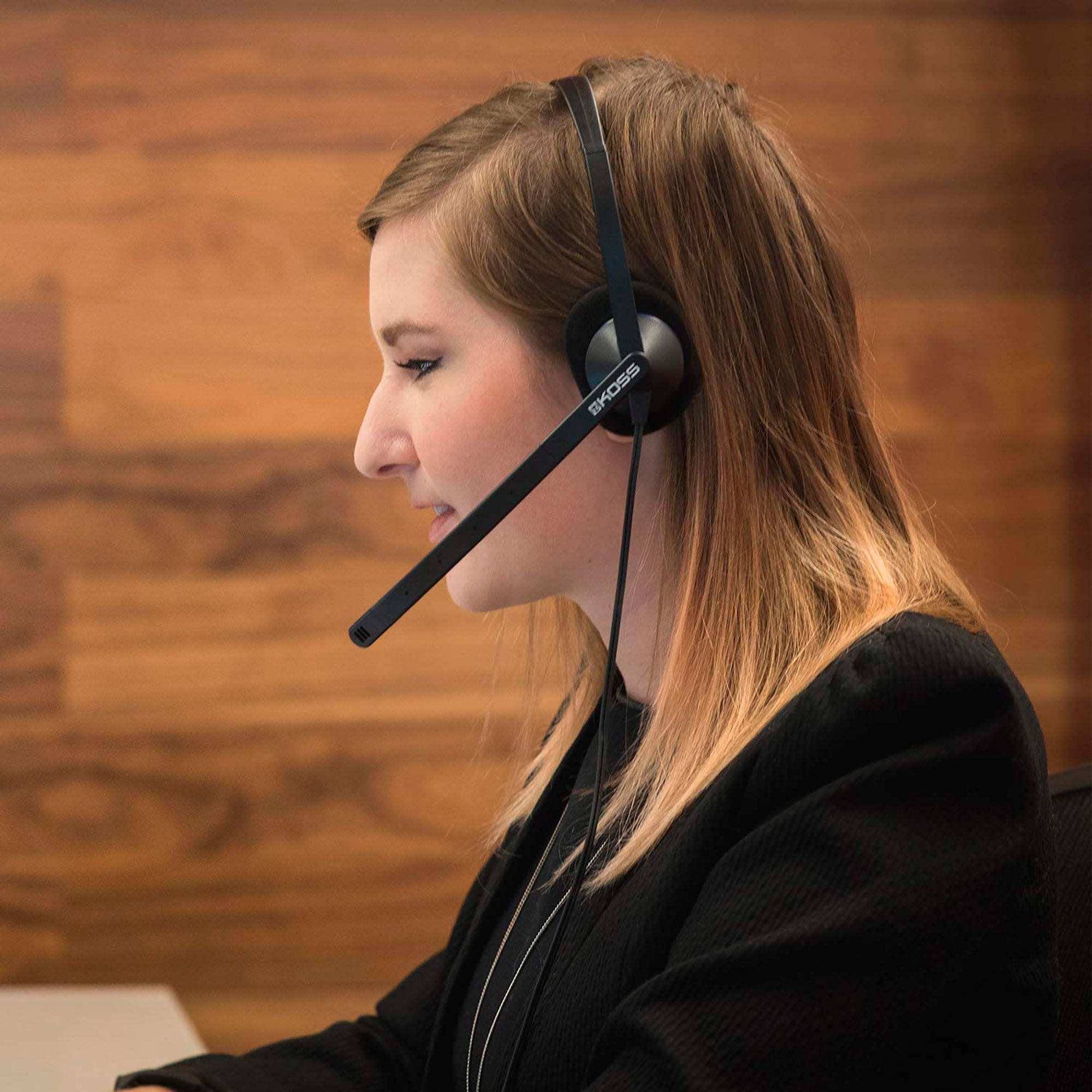 Kobieta w czarnych słuchawkach z mikrofonem, uśmiechnięta, w otoczeniu biurowym.