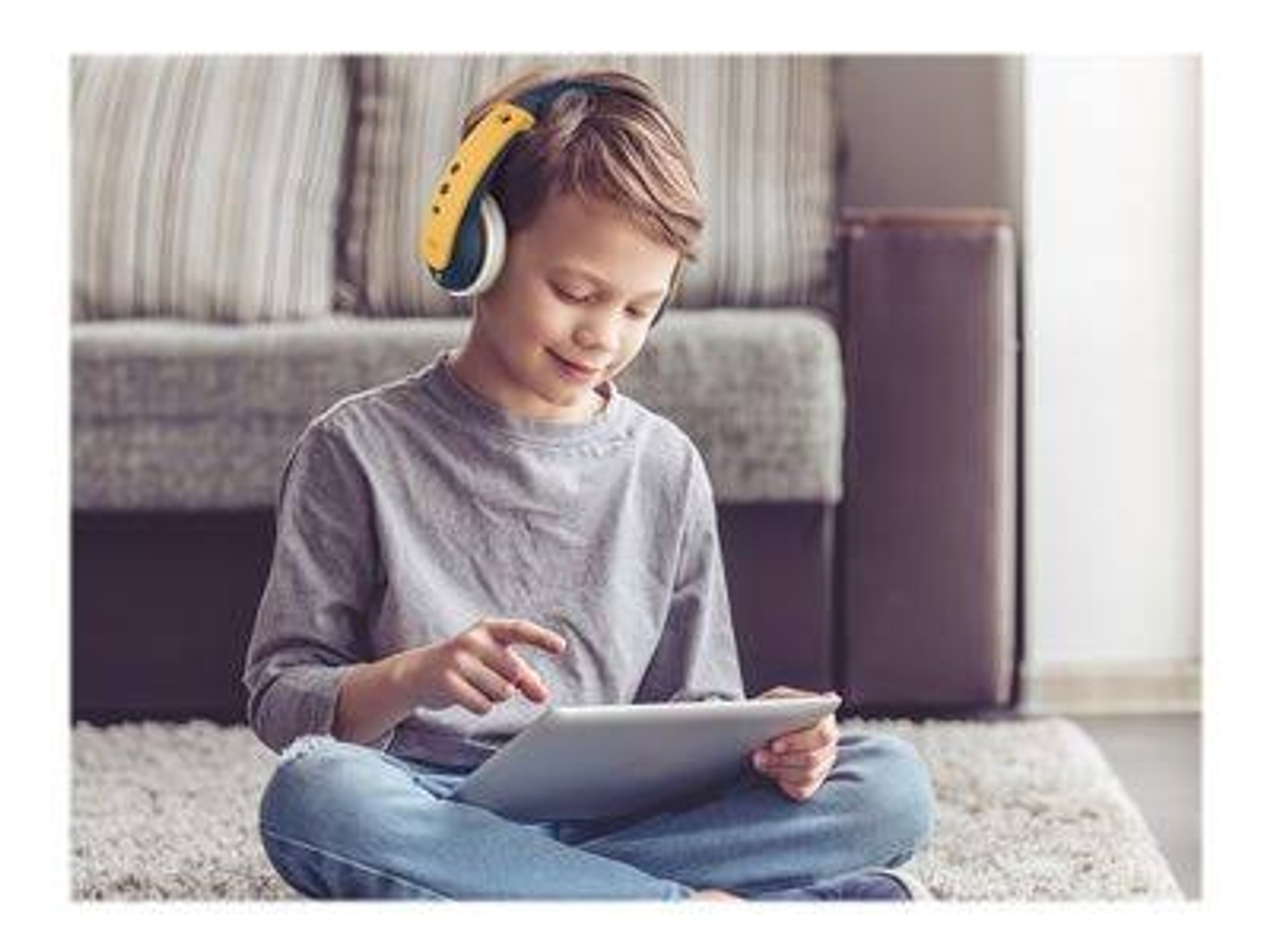 Un jeune garçon avec un casque est assis, utilisant une tablette. L'arrière-plan comprend un canapé et un tapis.