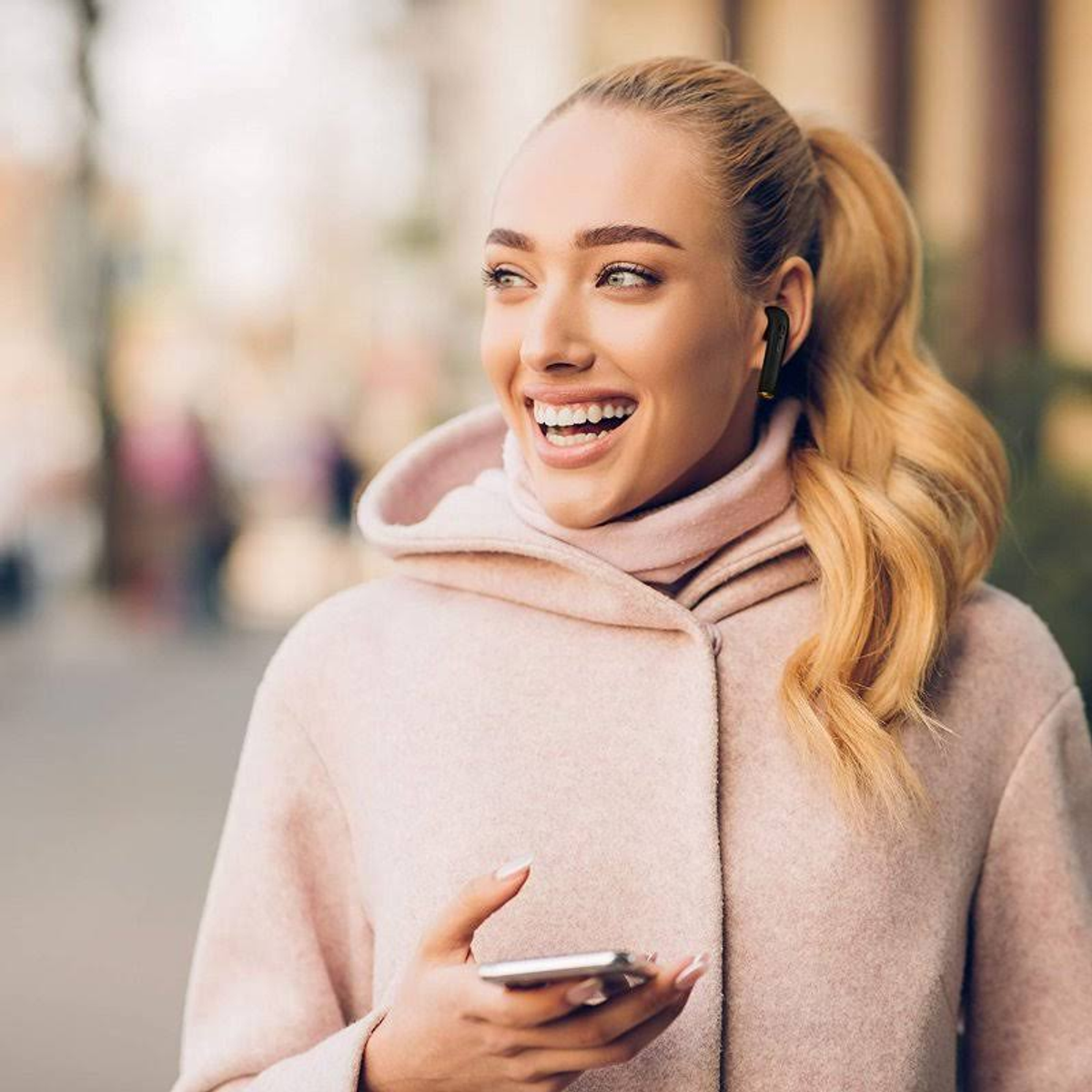 Donna con auricolari sorridente, tiene telefono. Capelli biondi, giacca rosa.