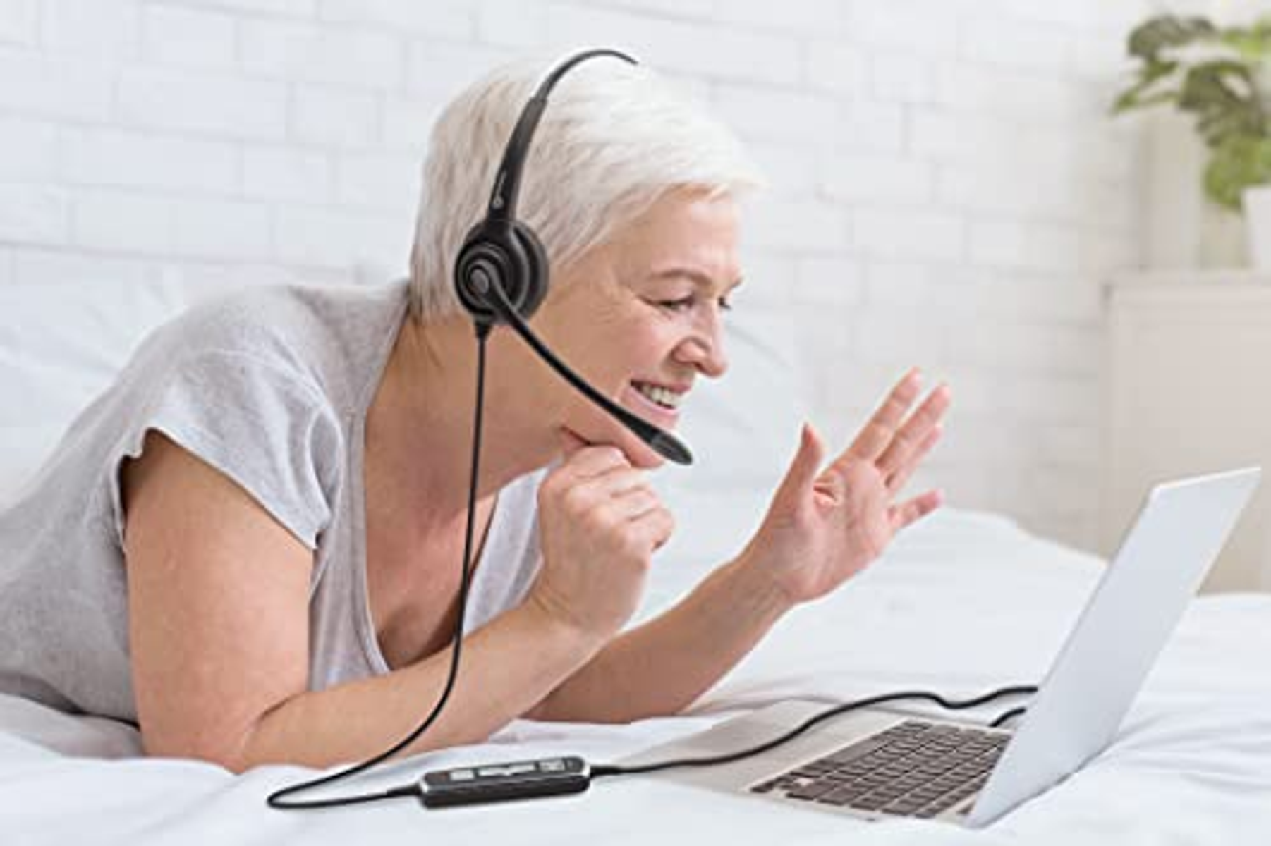 Starsza kobieta w zestawie słuchawkowym, uśmiechnięta i gestykulująca przed laptopem.