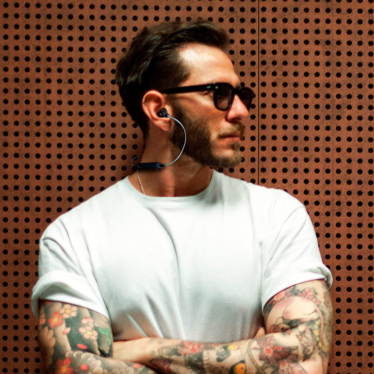 Uomo con auricolari e occhiali da sole, con tatuaggi, vicino a un muro.