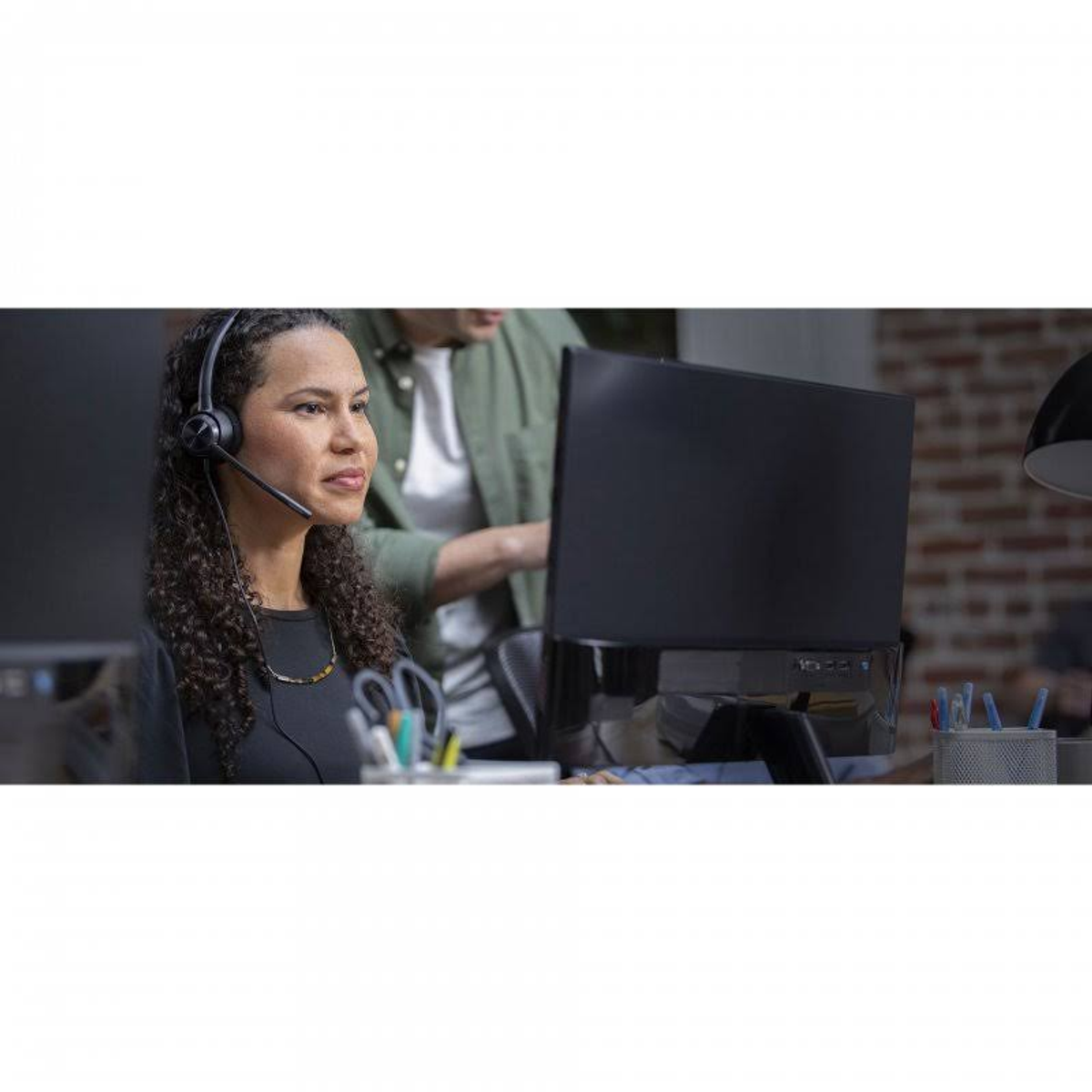 Eine Frau trägt ein Headset mit Mikrofon und arbeitet im Büro. Eine weitere Person im Hintergrund.