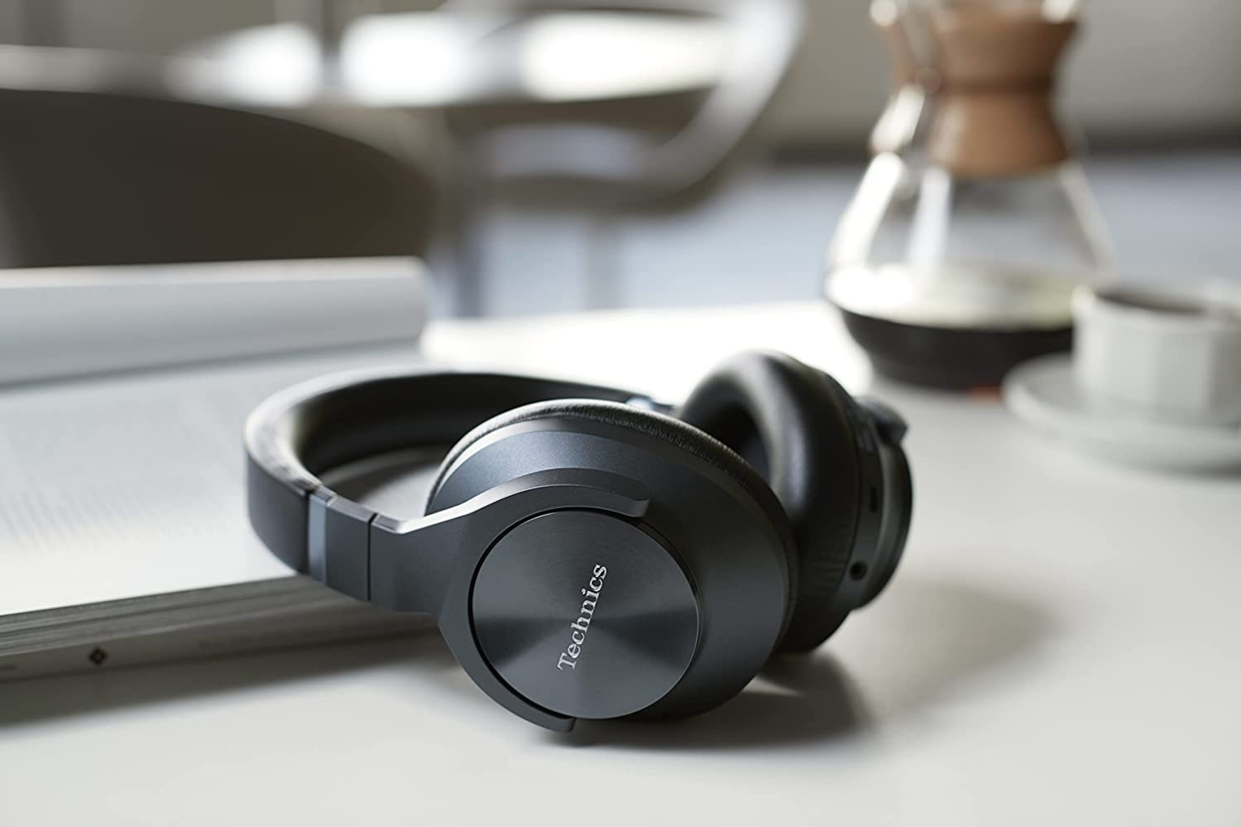 Black headphones resting on a white surface. A book and a cup are also on the table.