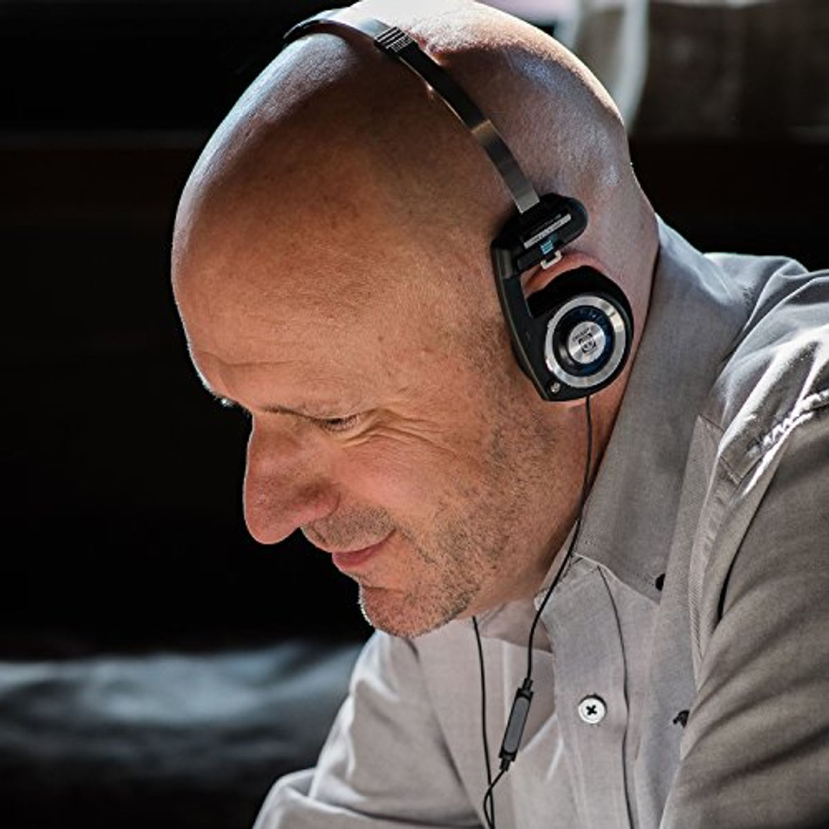 Łysy mężczyzna w czarnych słuchawkach. Patrzy w dół i uśmiecha się. Tło jest rozmyte.