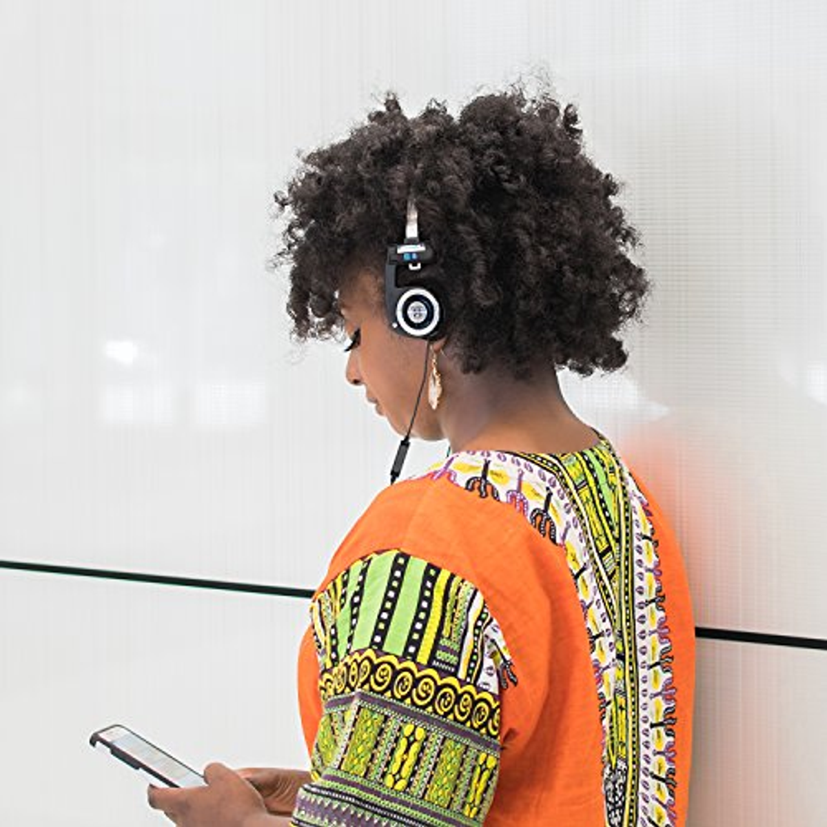 Kobieta z włosami afro w słuchawkach. Trzyma telefon. Tłem jest biała ściana.