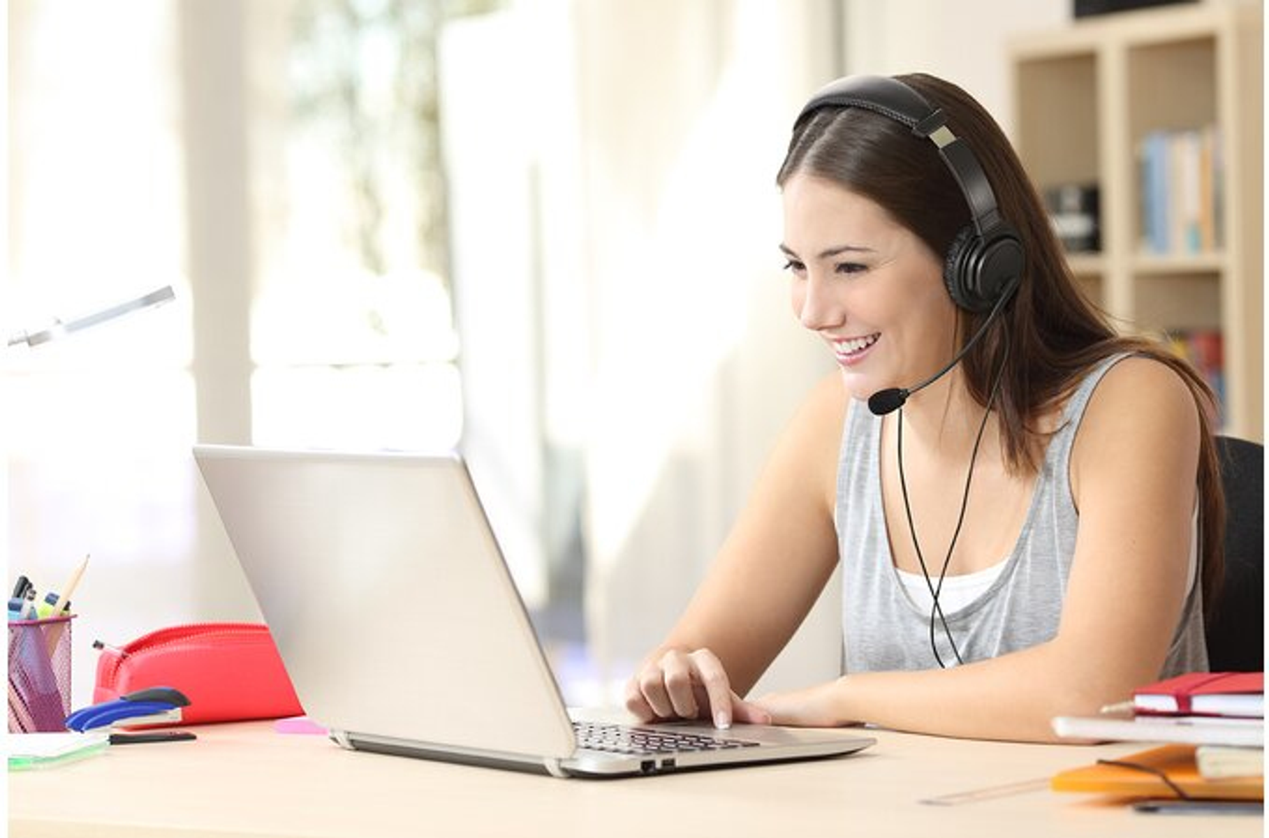 Vrouw met headset lacht naar laptop. Ze zit aan een bureau in een kamer met een raam.