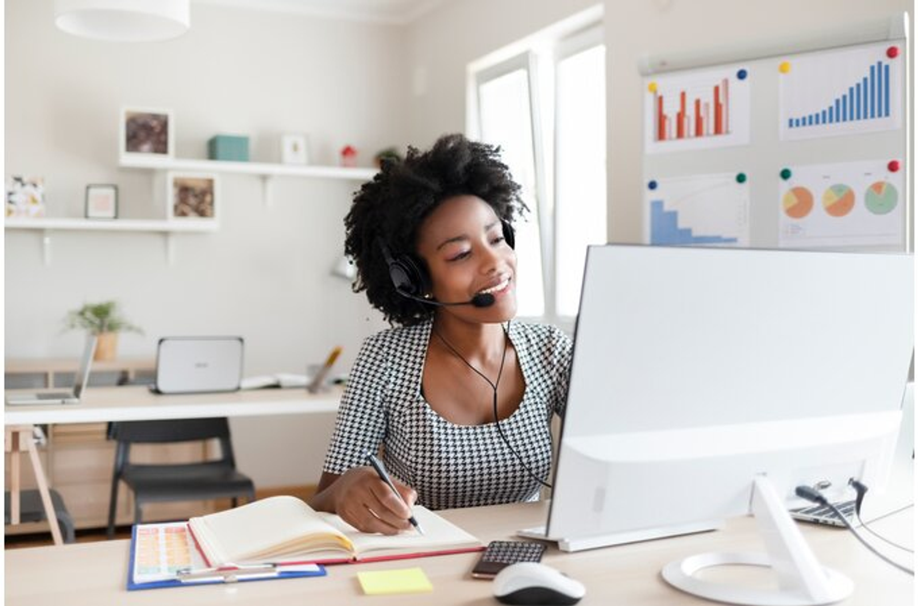Kobieta w zestawie słuchawkowym, uśmiechnięta, pracuje przy biurku z komputerem.