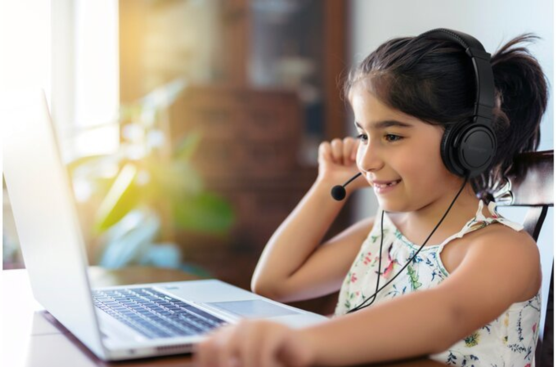 Dziewczyna w słuchawkach z mikrofonem uśmiecha się, patrząc na laptopa. Słońce świeci w tle.