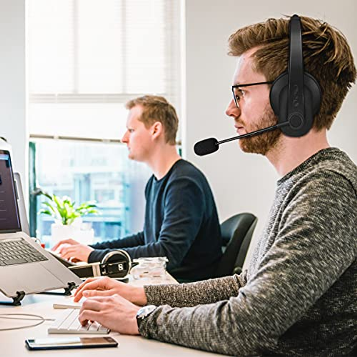 Dwóch mężczyzn używa laptopów. Jeden ma na sobie zestaw słuchawkowy. Otoczenie biurowe, białe tło.