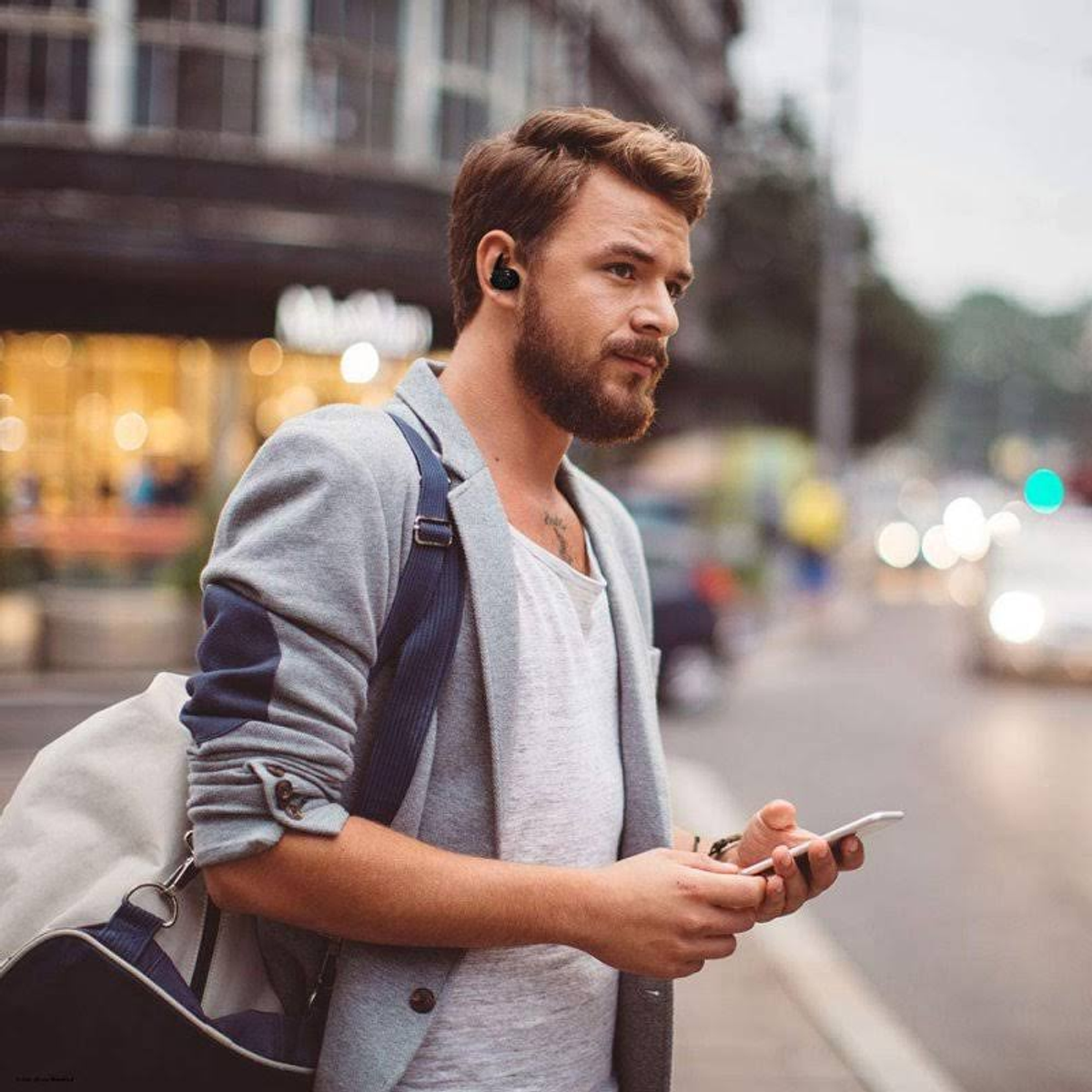 Un hombre usando auriculares, caminando por la ciudad, con una chaqueta gris y una bolsa.