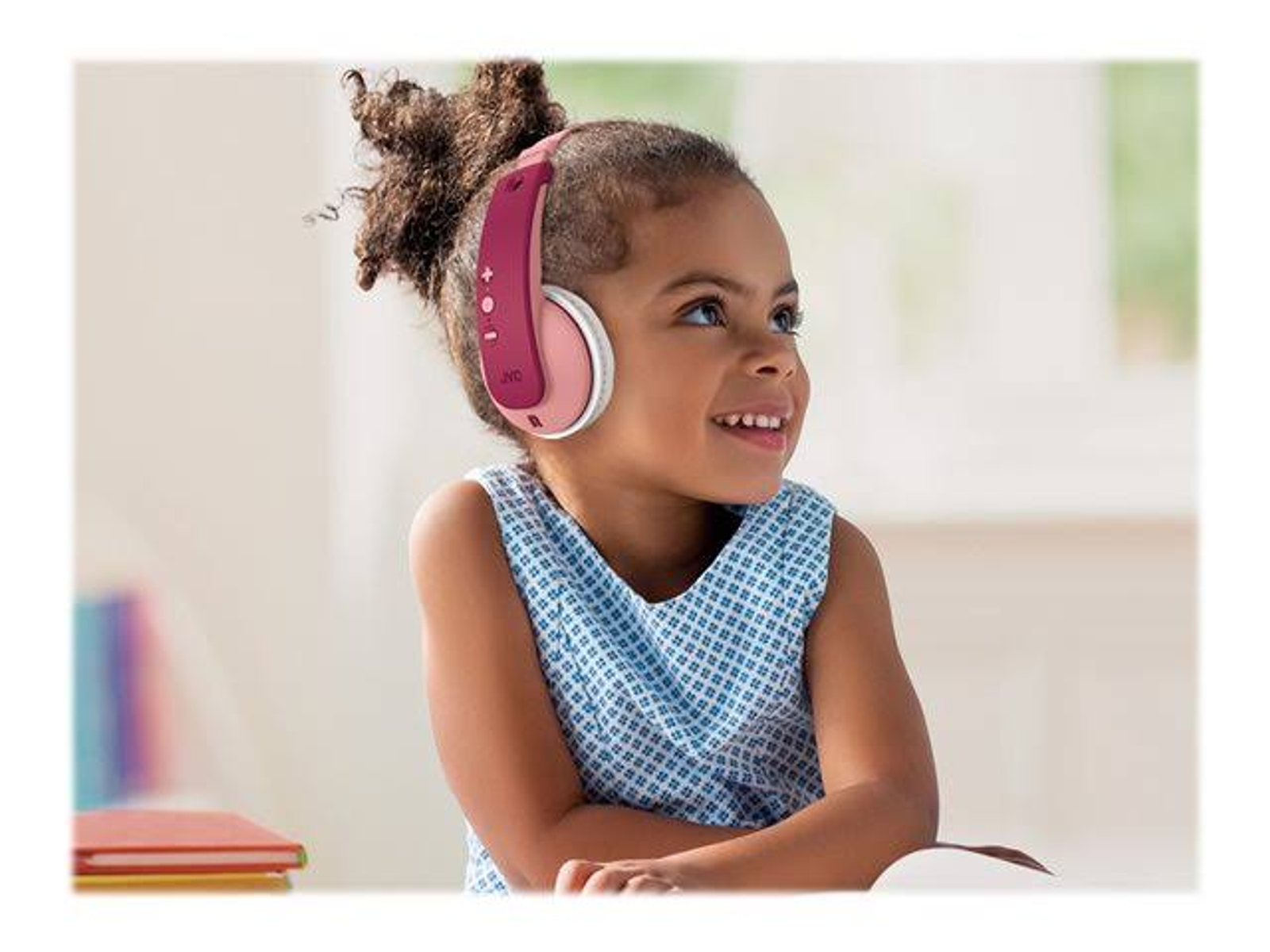 Una bambina sorridente con cuffie JVC rosa. Seduta in una stanza con libri su un tavolo.