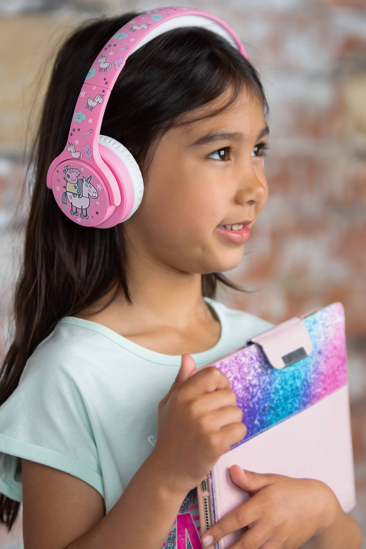 Niña con auriculares rosas sosteniendo una tableta. Una pared de ladrillos al fondo.