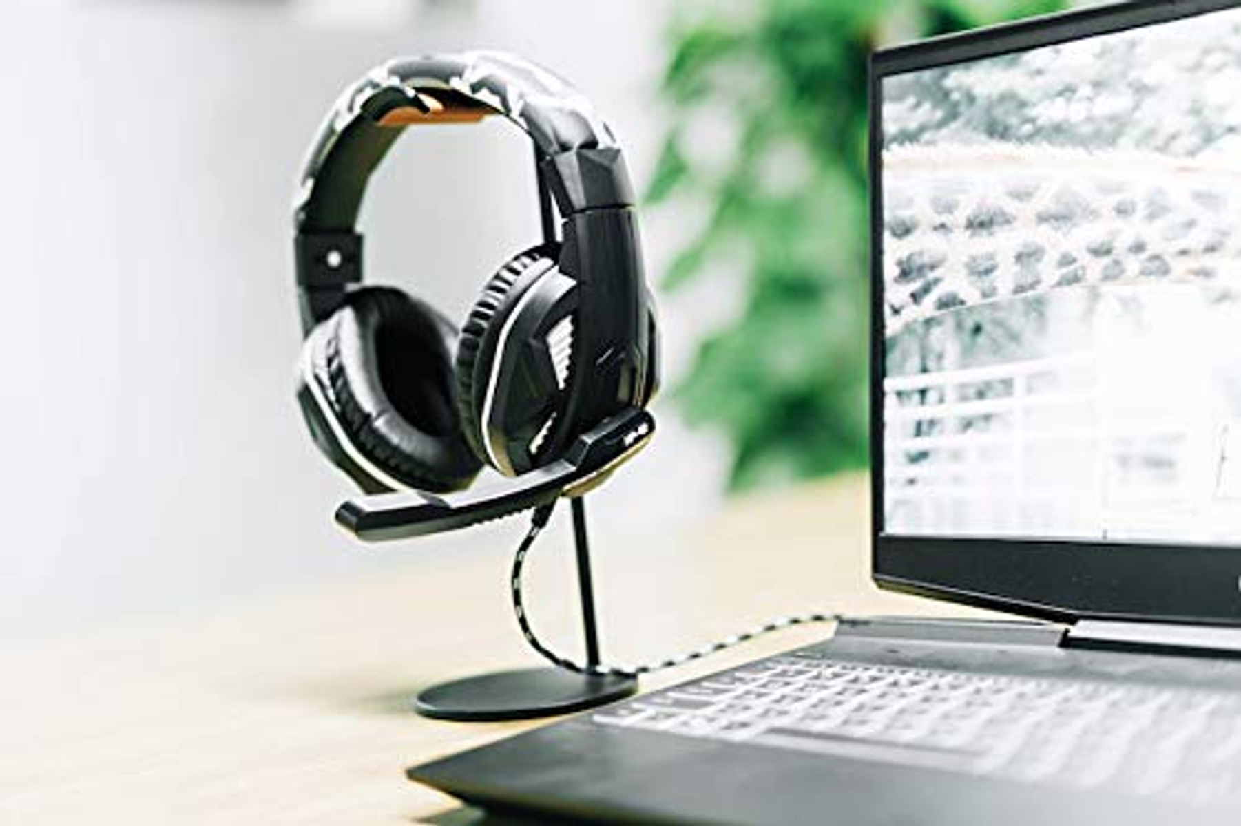 Czarno-białe słuchawki do gier na stojaku, obok laptopa na drewnianym biurku.
